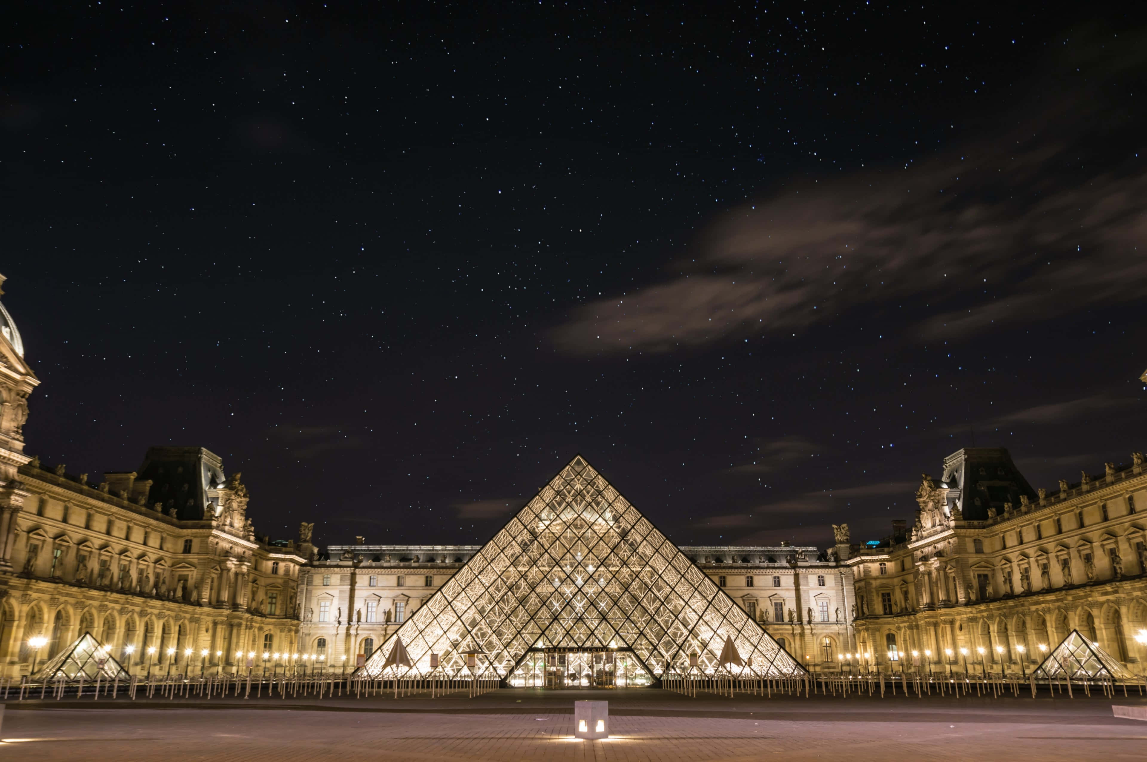french 桌布 壁紙 — 羅浮宮的玻璃金字塔在繁星點點的夜空下發光，薄雲飄浮在其歷史悠久的石翼上方。
