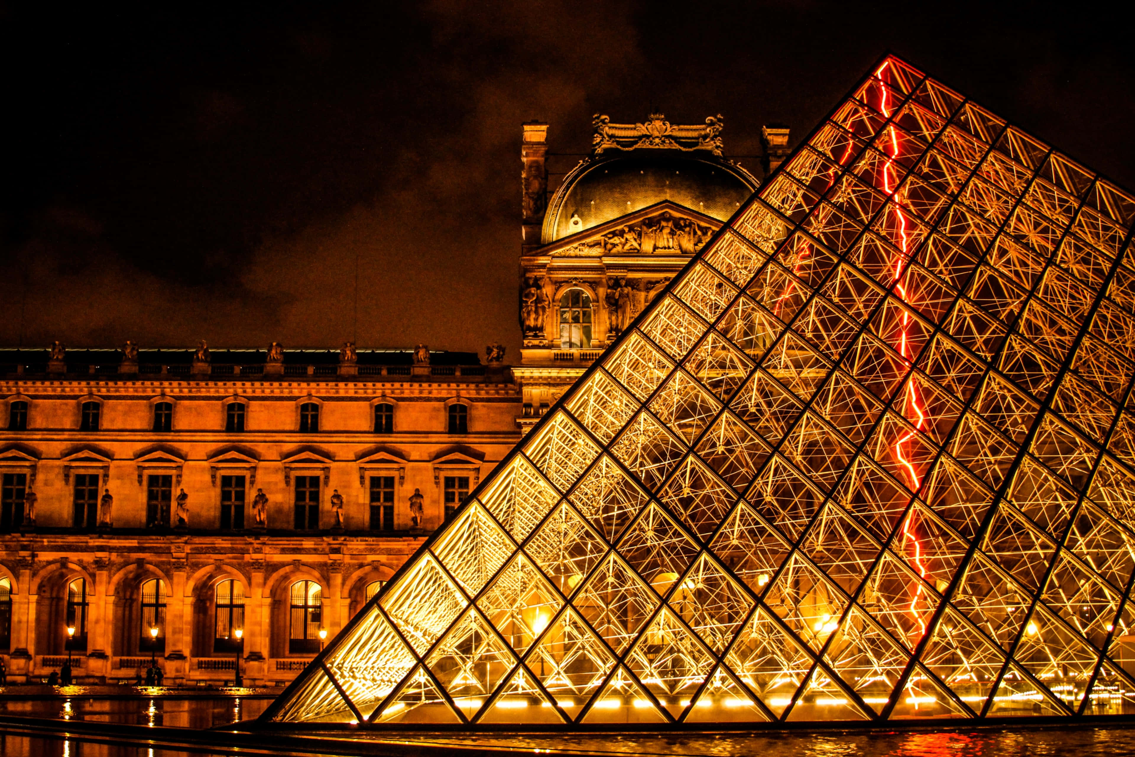 France 桌布 壁紙 — 羅浮宮金字塔在夜晚溫暖地發光，紅色閃電照亮其玻璃結構，映襯著歷史悠久的宮殿外牆。