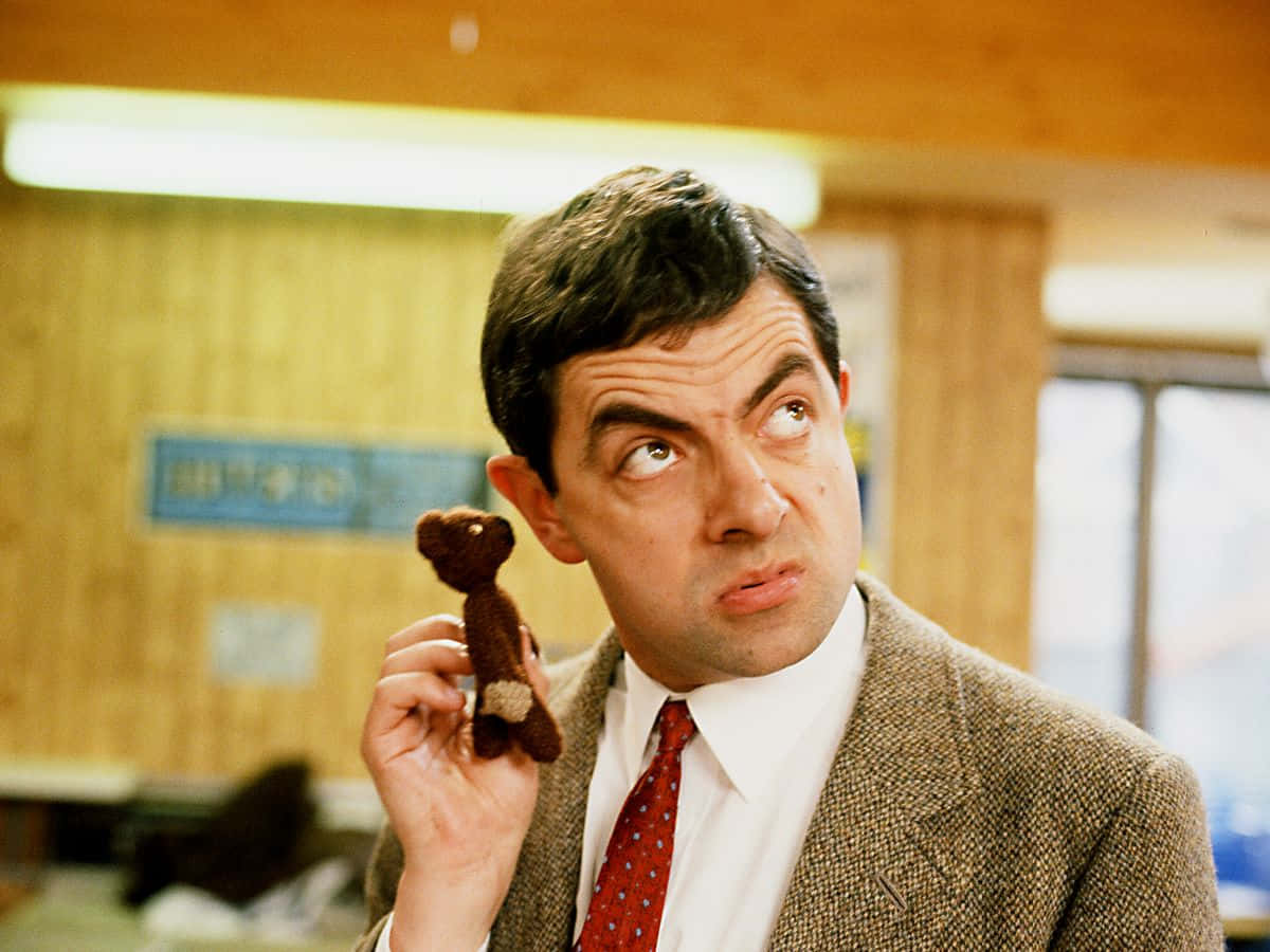 豆豆先生 桌布 壁紙 — 羅溫·艾金森飾演Mr Bean，在辦公室裡抱著一隻小棕色泰迪熊，露出滑稽而懷疑的表情。