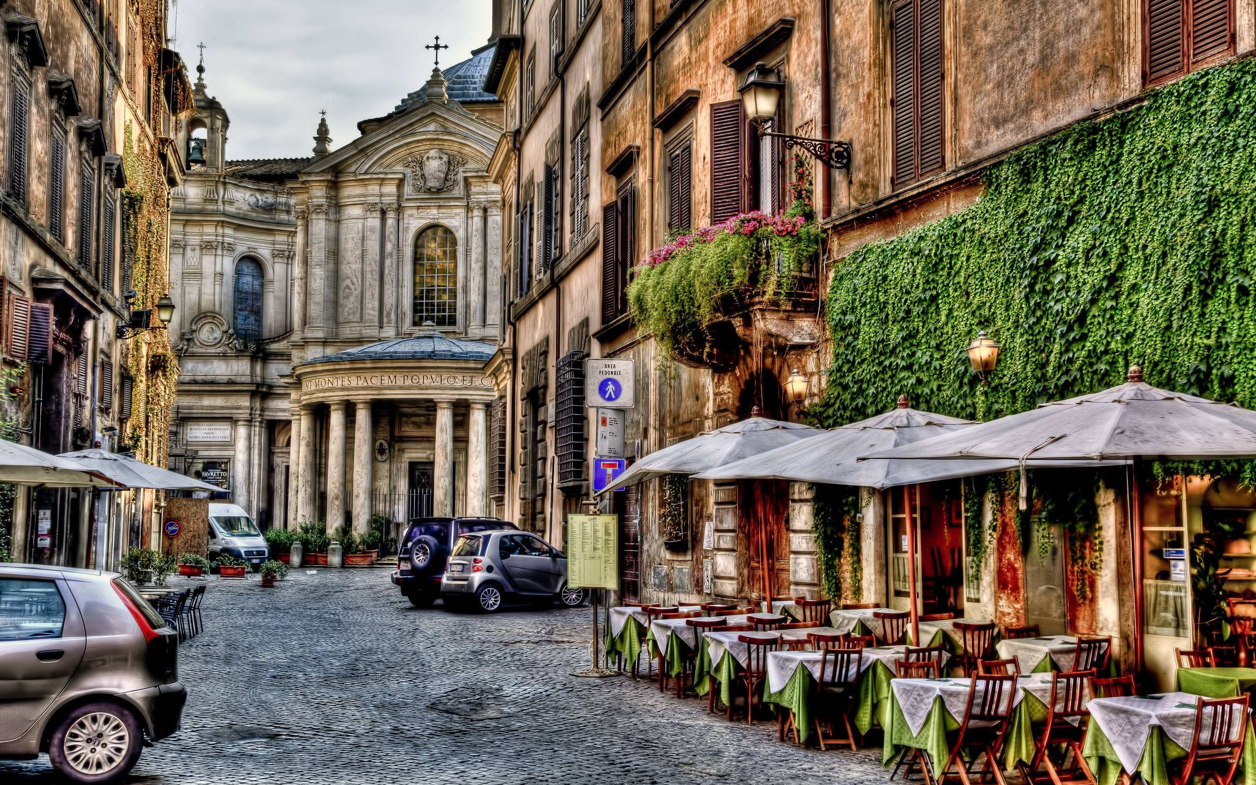Rome 桌布 壁紙 — 羅馬一條狹窄的鵝卵石街道，兩旁是爬滿常春藤的建築物，遮陽傘下設有戶外咖啡桌。