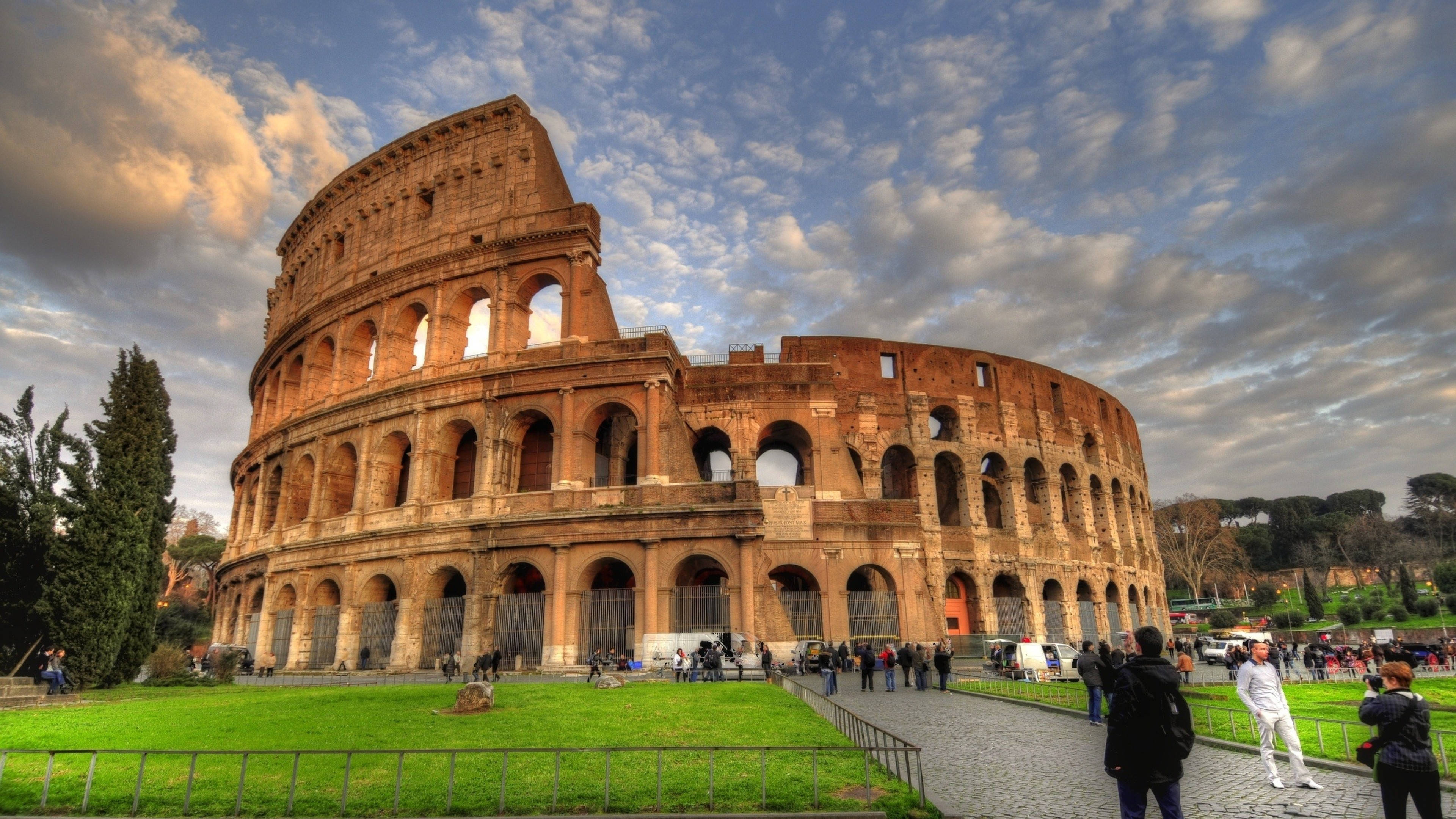 Rome 桌布 壁紙 — 羅馬的古代競技場在戲劇性的多雲天空下雄偉地矗立，遊客正在探索其歷史遺跡。