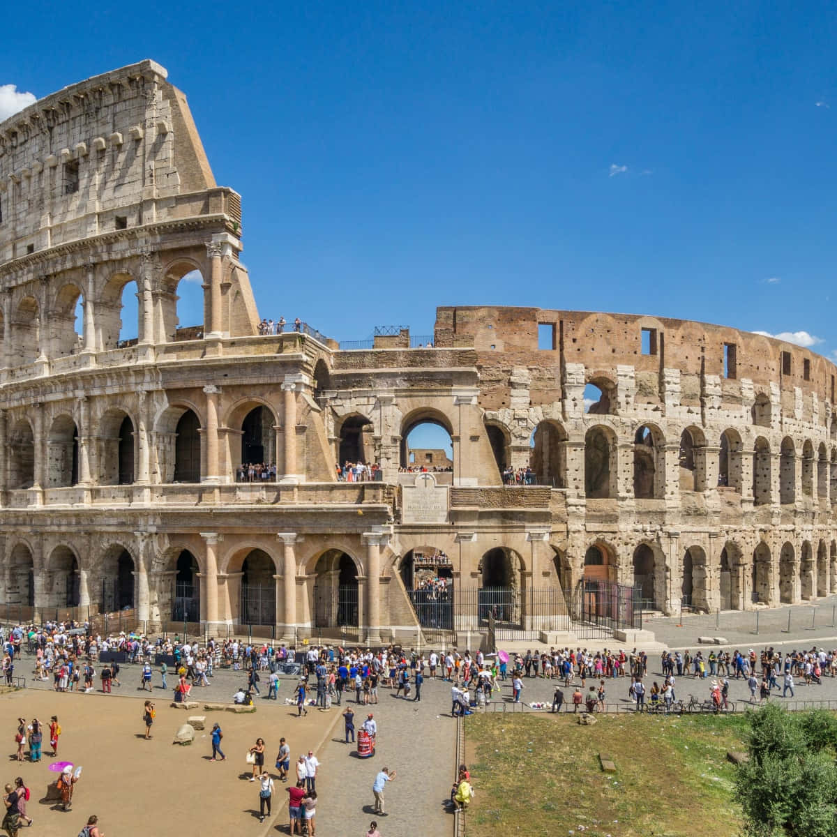 Roman Colosseum 桌布 壁紙 — 羅馬的古代競技場在晴朗的藍天下雄偉地矗立，成群結隊的遊客正在探索其歷史遺跡。