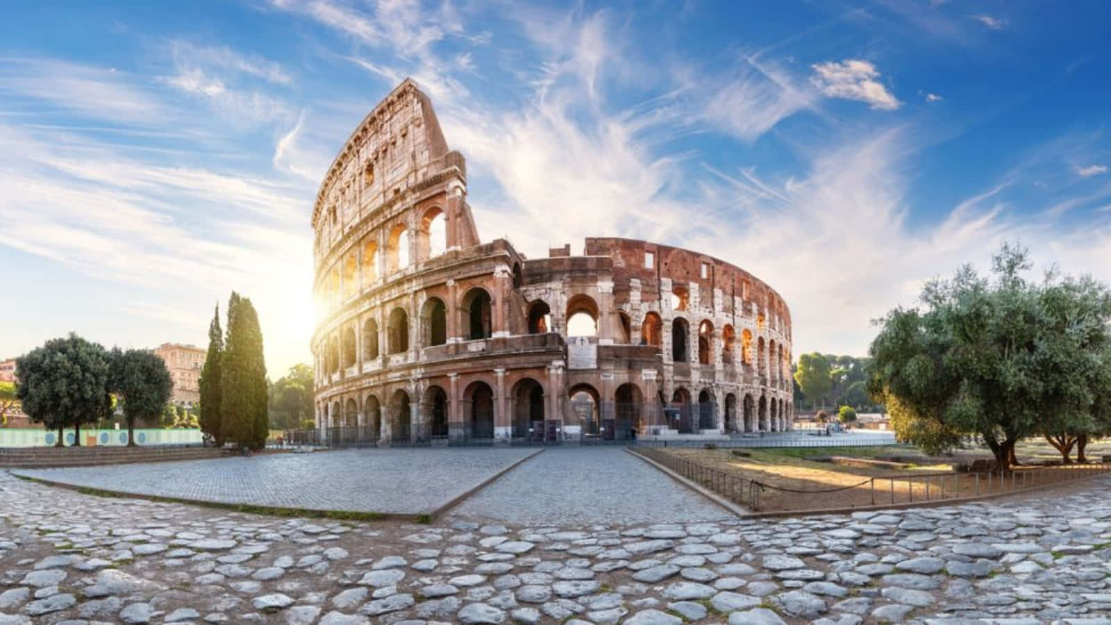 Roman 桌布 壁紙 — 羅馬的古代競技場沐浴在金色的日出光芒中，鵝卵石小徑和綠樹環繞著這座歷史遺跡。