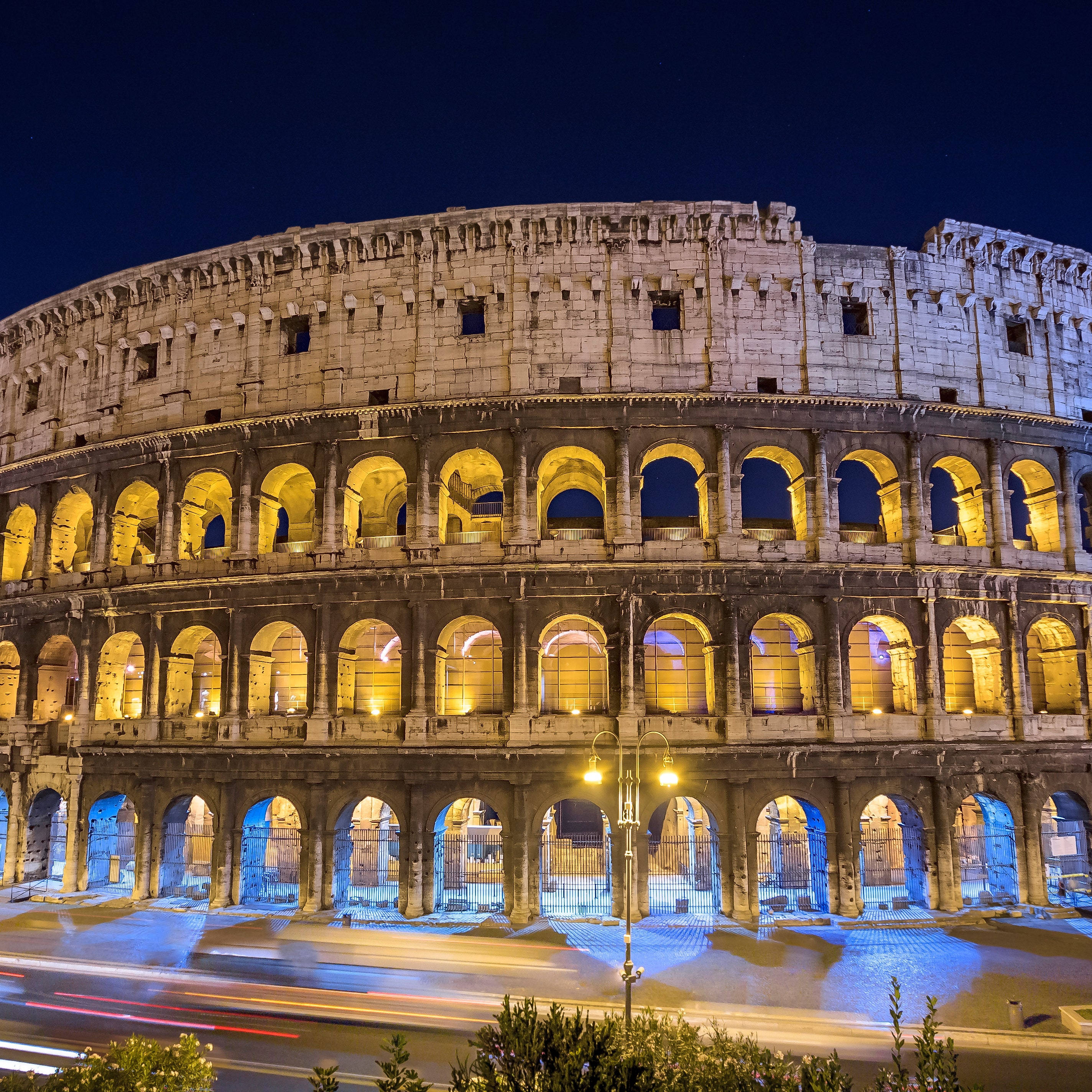 Rome 桌布 壁紙 — 羅馬競技場在夜燈下溫暖地發光，帶有藍色點綴和下方模糊的車流痕跡。