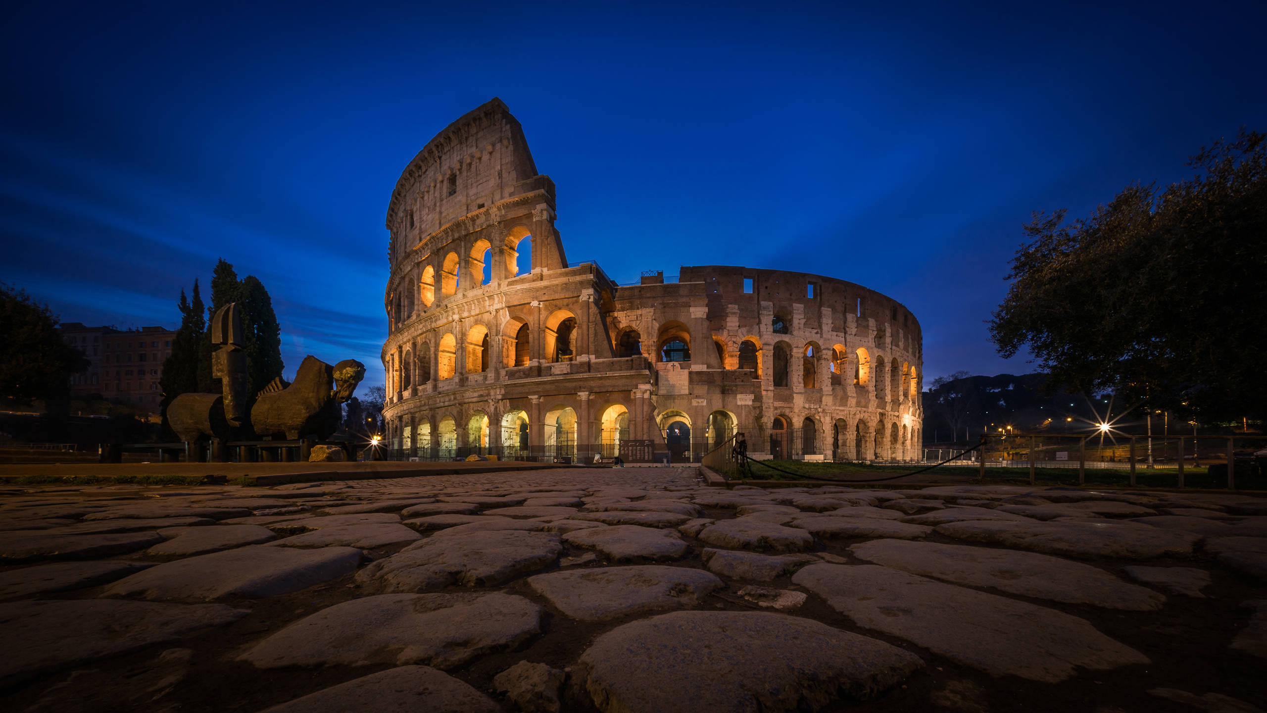 Rome 桌布 壁紙 — 羅馬競技場在暮色天空下溫暖地發光，鵝卵石小徑通往其古老的拱門。