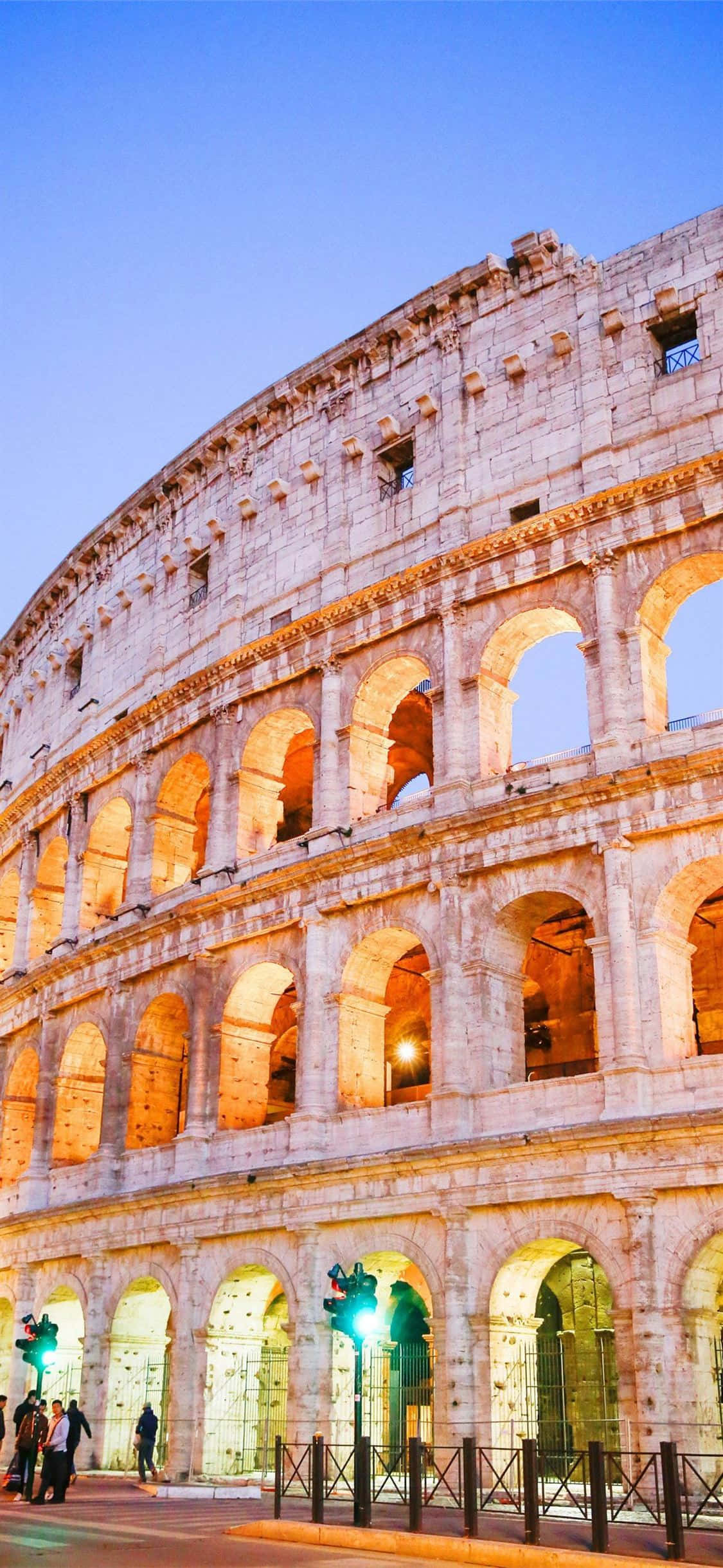 Italy Aesthetic 桌布 壁紙 — 羅馬競技場在暮色燈光下溫暖地發光，襯托著清澈的藍天，遊客在附近漫步。