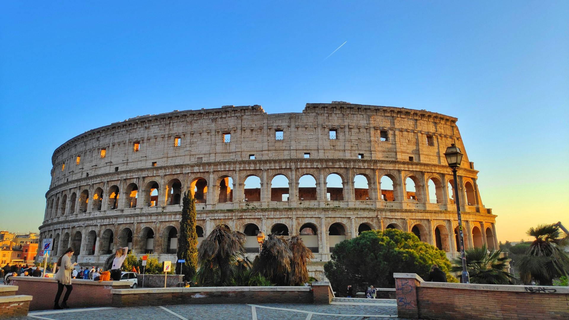 Rome 桌布 壁紙 — 羅馬競技場沐浴在金色夕陽下，附近有遊客走動，前景有棕櫚樹。