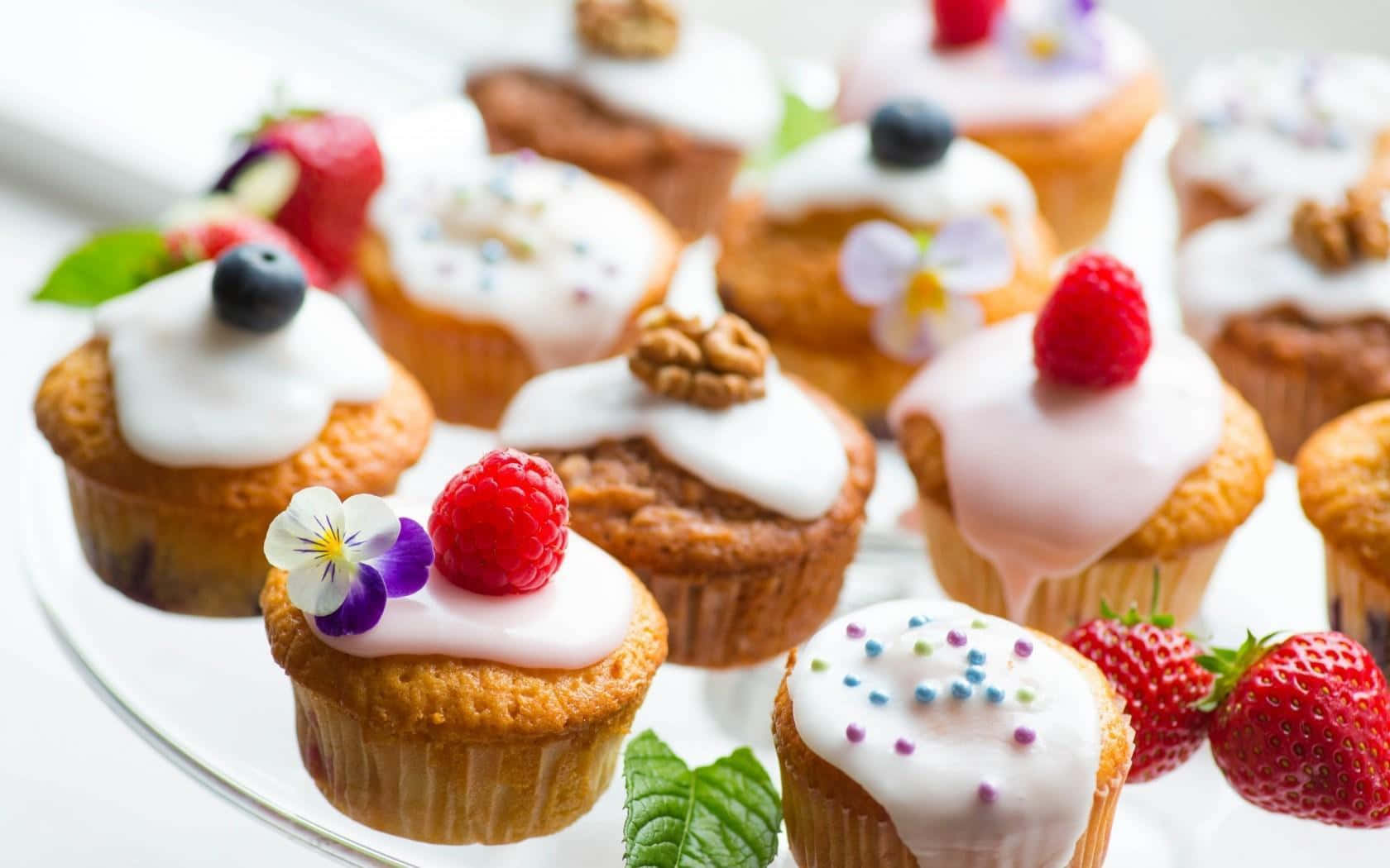 Desserts 桌布 壁紙 — 美味的杯子蛋糕，上面有白色糖霜、新鮮漿果和可食用花卉，擺放在玻璃盤上。