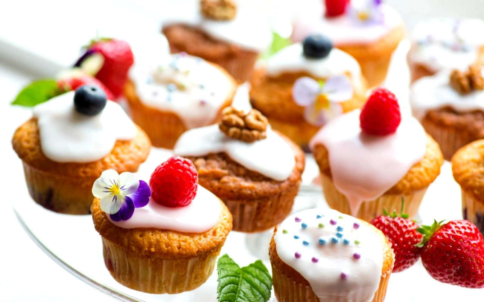 Spring Desserts 桌布 壁紙 — 美味的杯子蛋糕，上面有白色糖霜、新鮮莓果和食用花，擺在玻璃盤上。