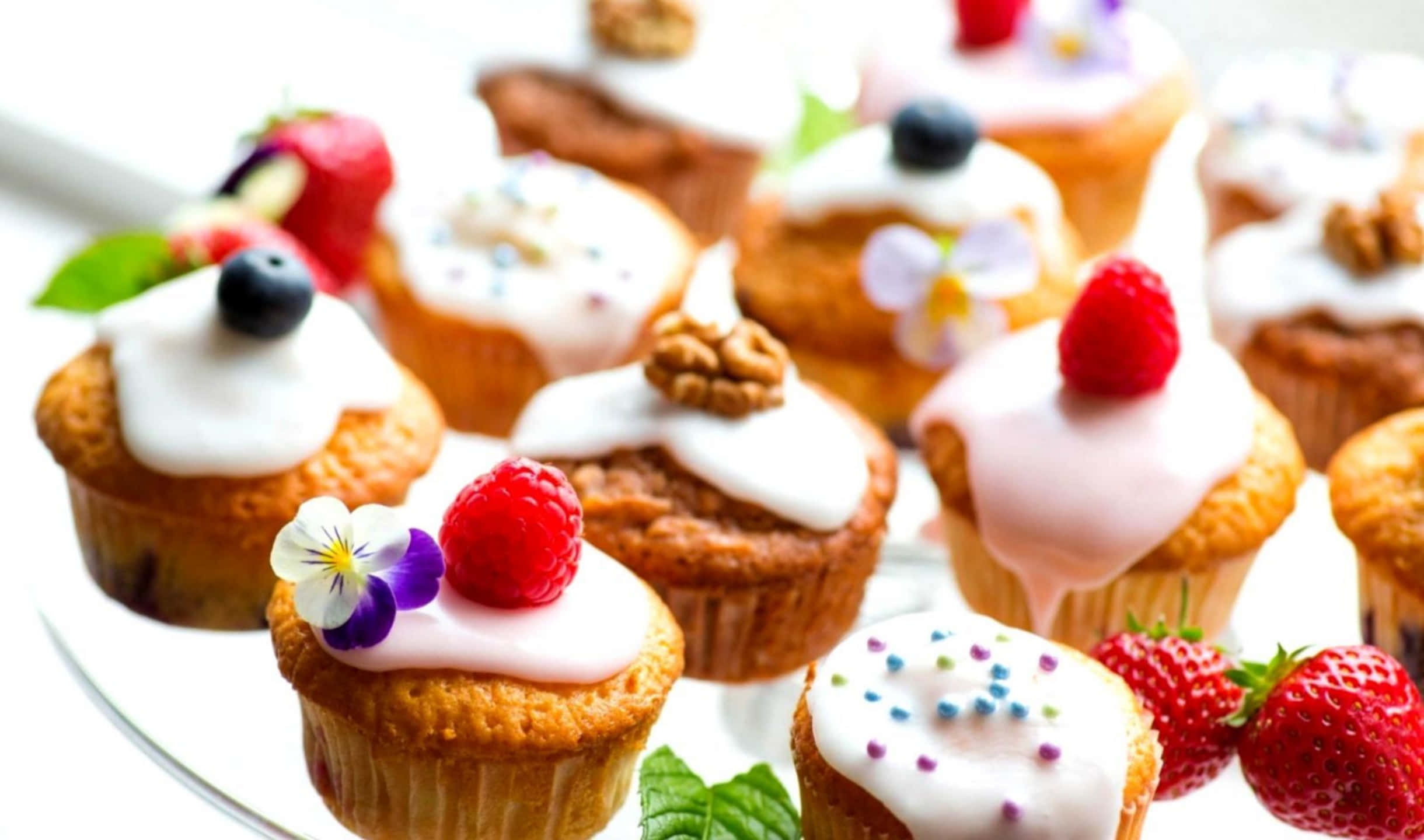 2440x1440 Desserts 桌布 壁紙 — 美味的杯子蛋糕，上面鋪滿白色糖霜、新鮮莓果和食用花，整齊擺放在玻璃盤上。