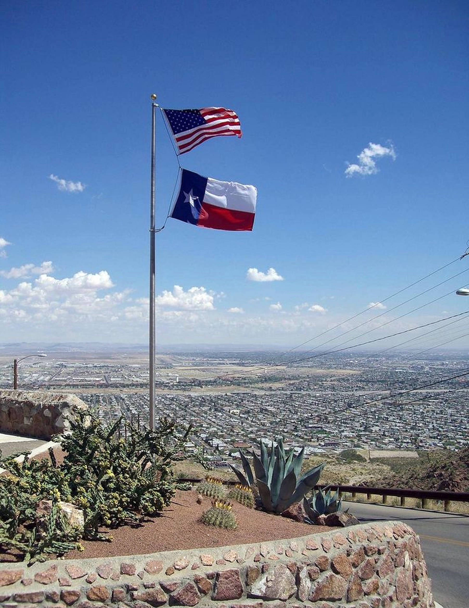 Texas Flag 桌布 壁紙 — 美國和德克薩斯州的旗幟在桿頂飄揚，俯瞰著廣闊的沙漠城市，天空湛藍，雲朵稀疏。