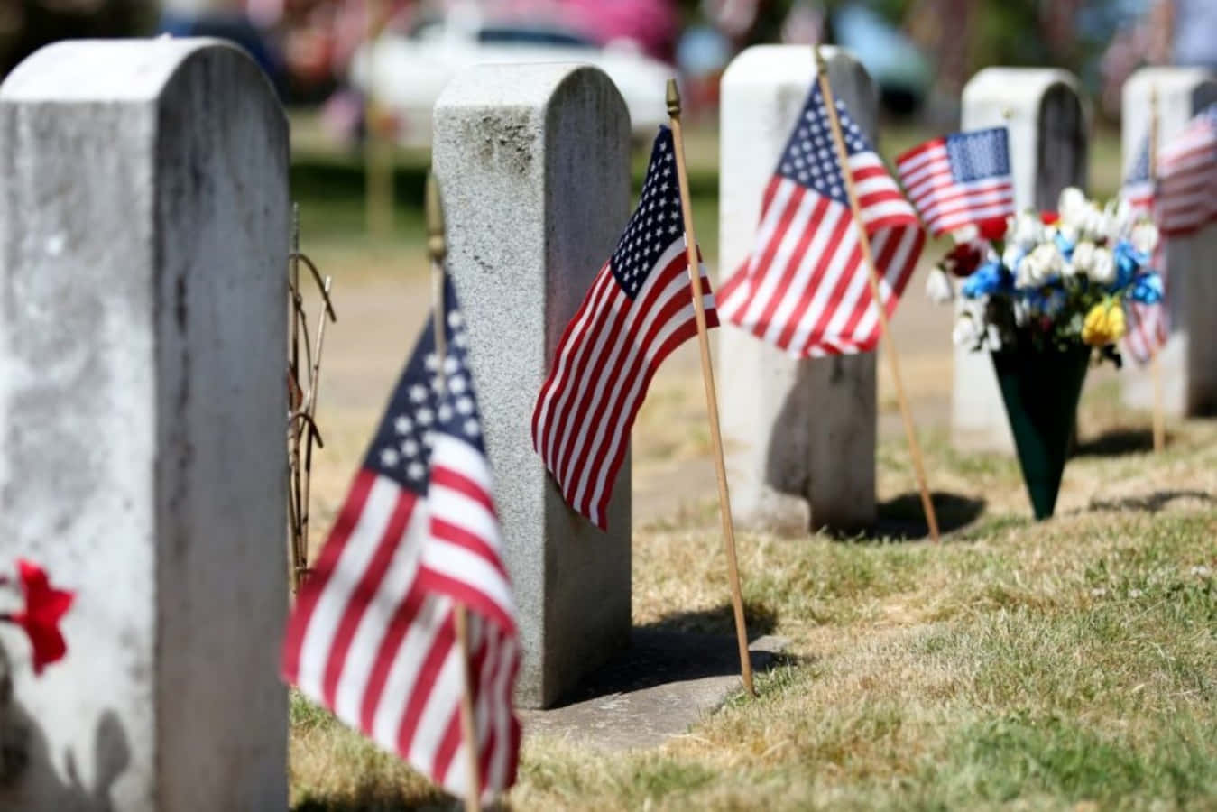 Memorial Day 桌布 壁紙 — 美國國旗和鮮花祭品點綴著陽光普照的墓園中風化的墓碑，天空晴朗。