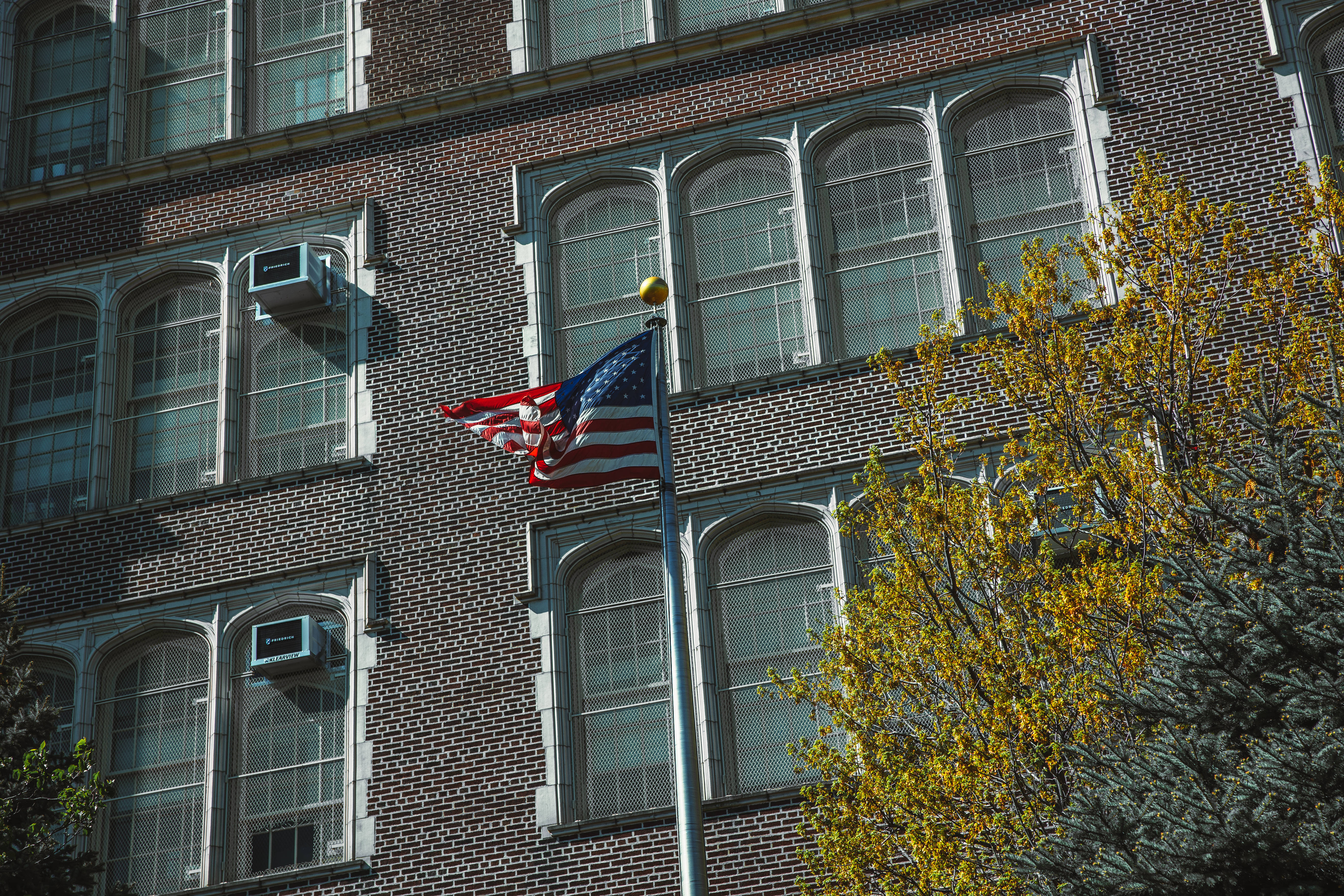 USA flag iPhone 桌布 壁紙 — 美國國旗在帶有拱形窗戶和秋樹的磚砌建築前自豪地飄揚。
