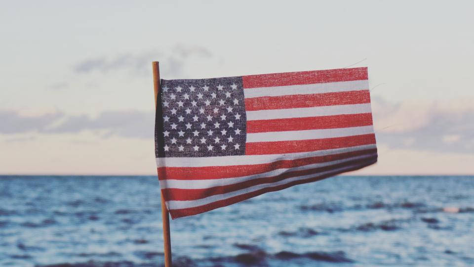 USA flag iPhone 桌布 壁紙 — 美國國旗在平靜的海洋地平線上空，對著柔和的暮色天空自豪地飄揚。