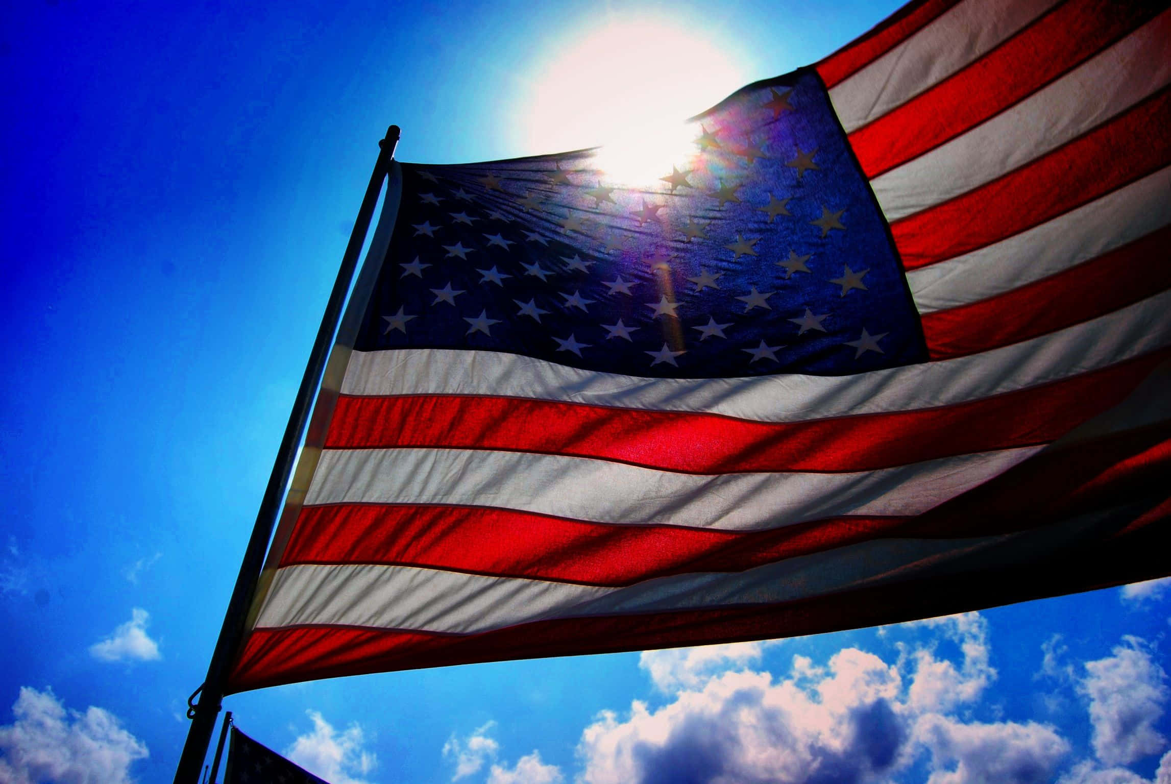 Veterans Day 桌布 壁紙 — 美國國旗在明亮的藍天下自豪地飄揚，陽光穿過其星星和條紋。