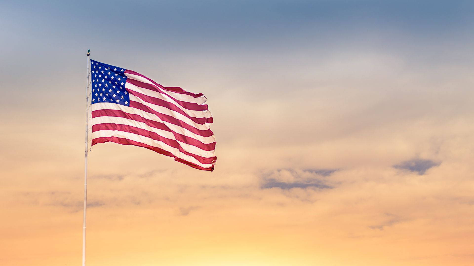 Flag 桌布 壁紙 — 美國國旗在柔和的金色日落天空和散落的雲朵中自豪地飄揚。