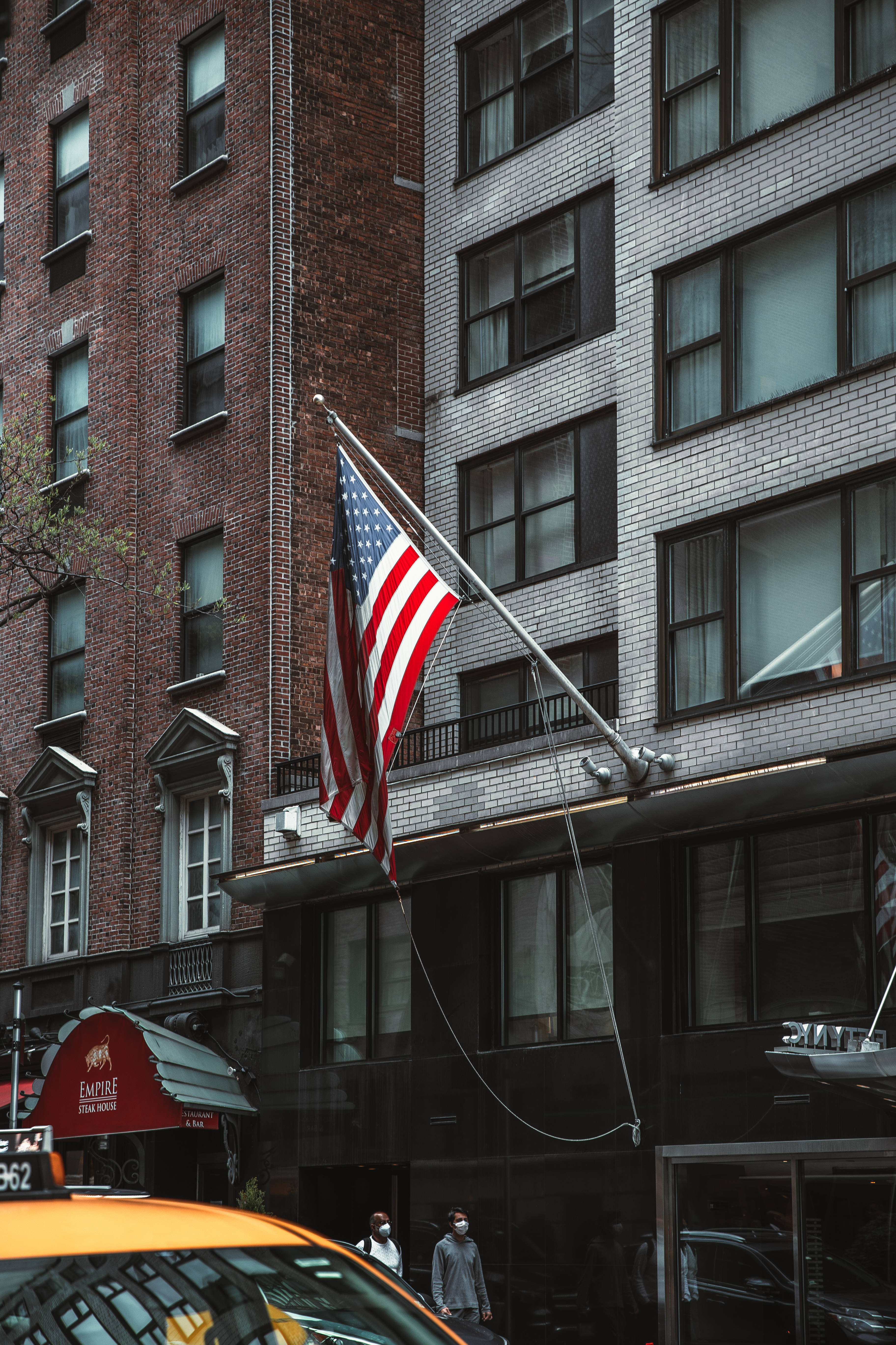 American Flag iPhone 桌布 壁紙 — 美國國旗在紅磚與白磚建築之間自豪地飄揚，下方有一輛黃色計程車駛過。