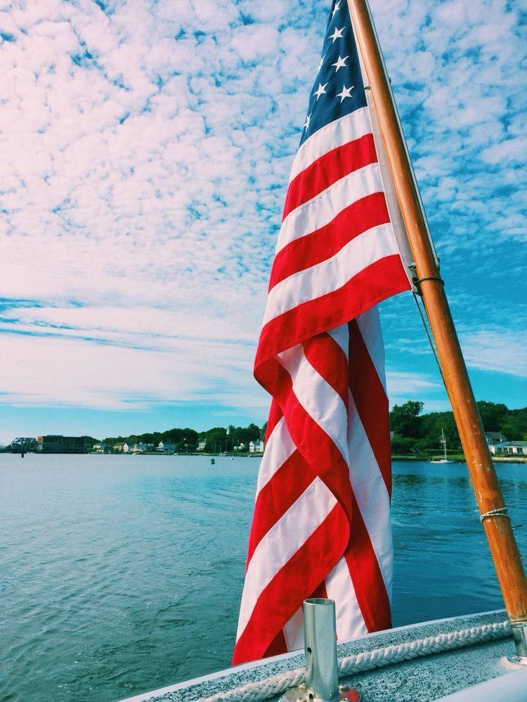 fourth of july 桌布 壁紙 — 美國國旗在船桅上飄揚，背景是明亮的藍天、散落的雲朵和平靜的水面。