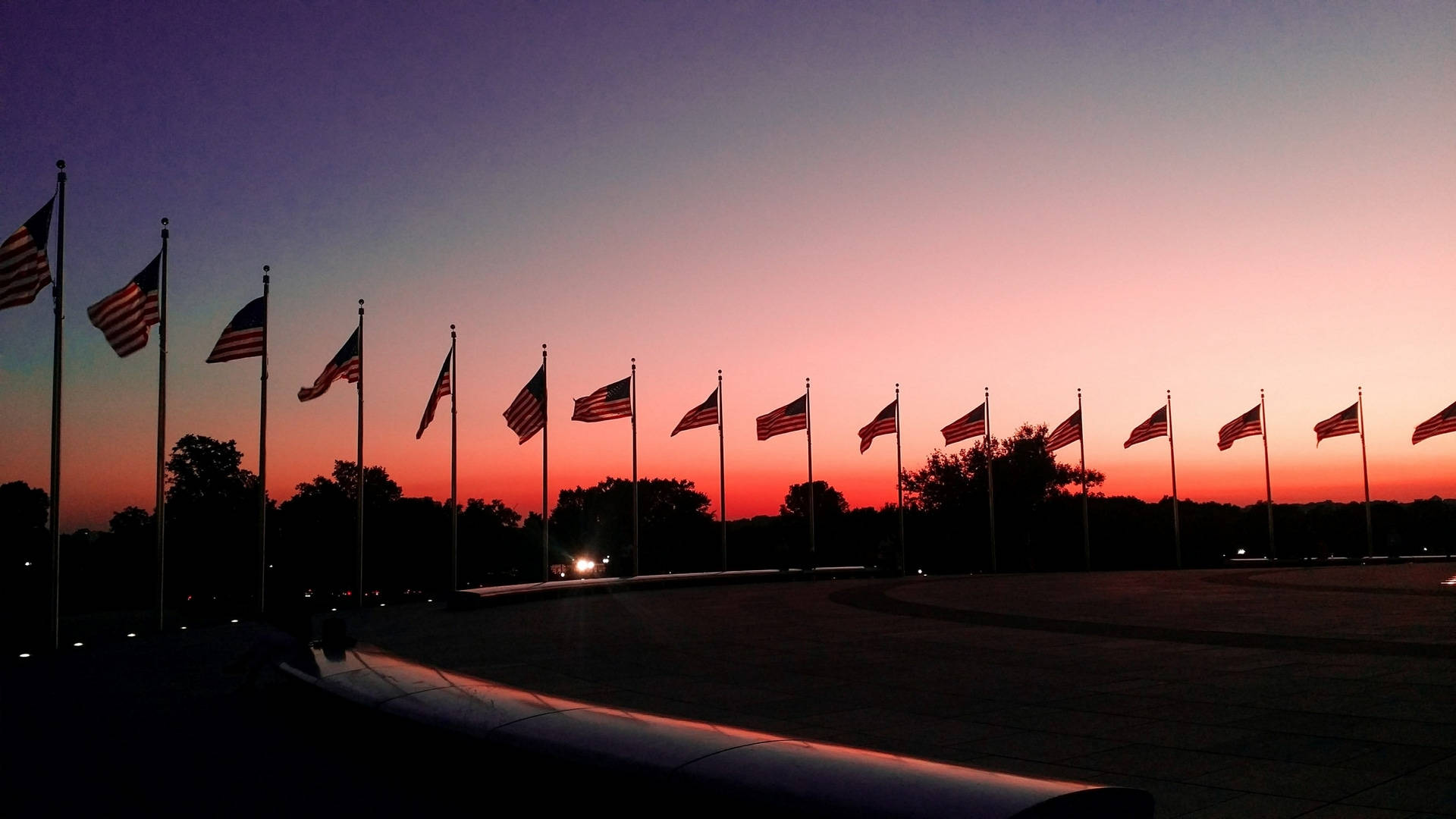 Flag 桌布 壁紙 — 美國國旗的剪影排列在廣場上，天空閃耀著橙色和紫色的日落色調。