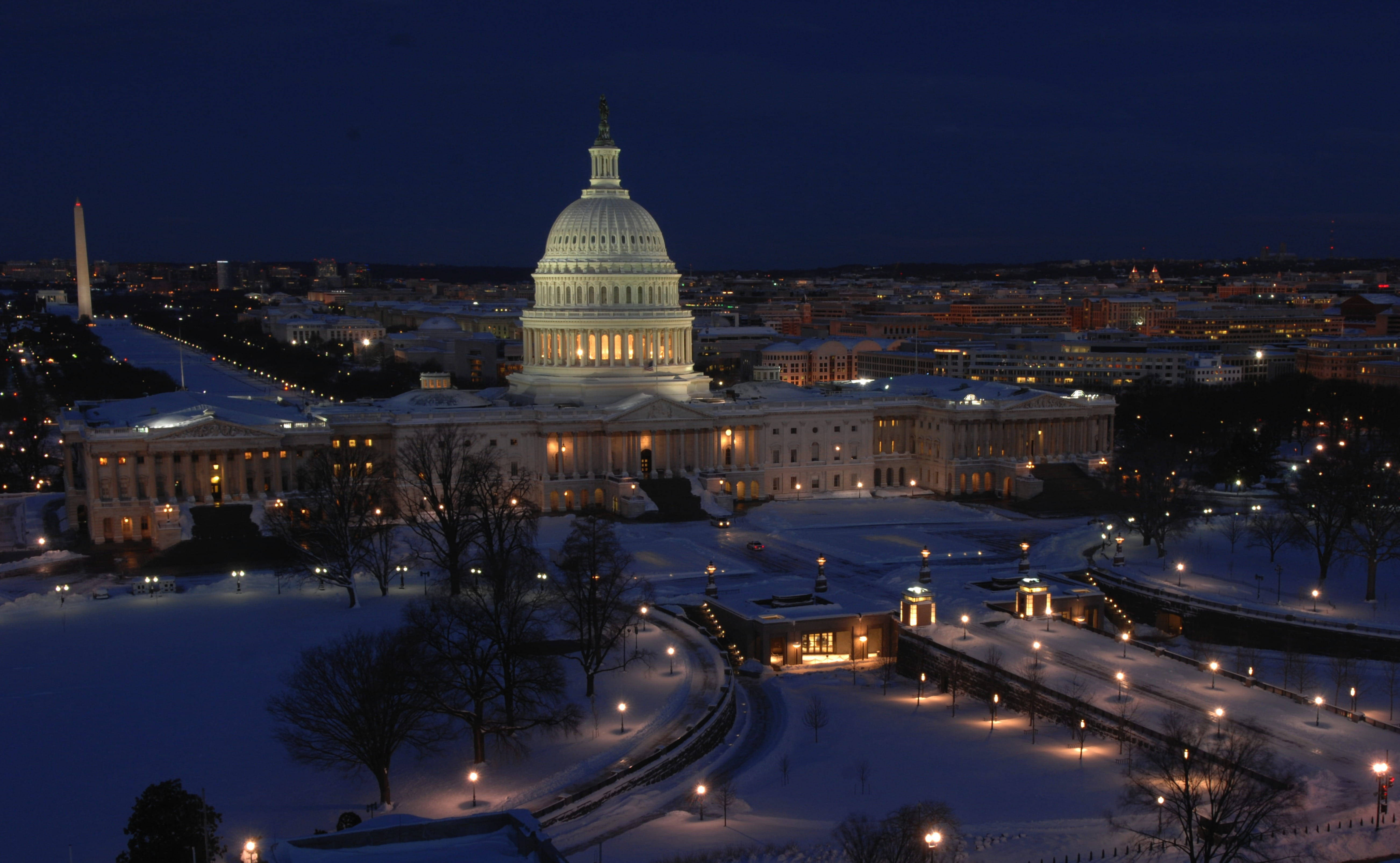 Washington 桌布 壁紙 — 美國國會大廈在夜晚雪覆蓋的地面上溫暖地發光，周圍環繞著城市燈光和光禿禿的冬季樹木。