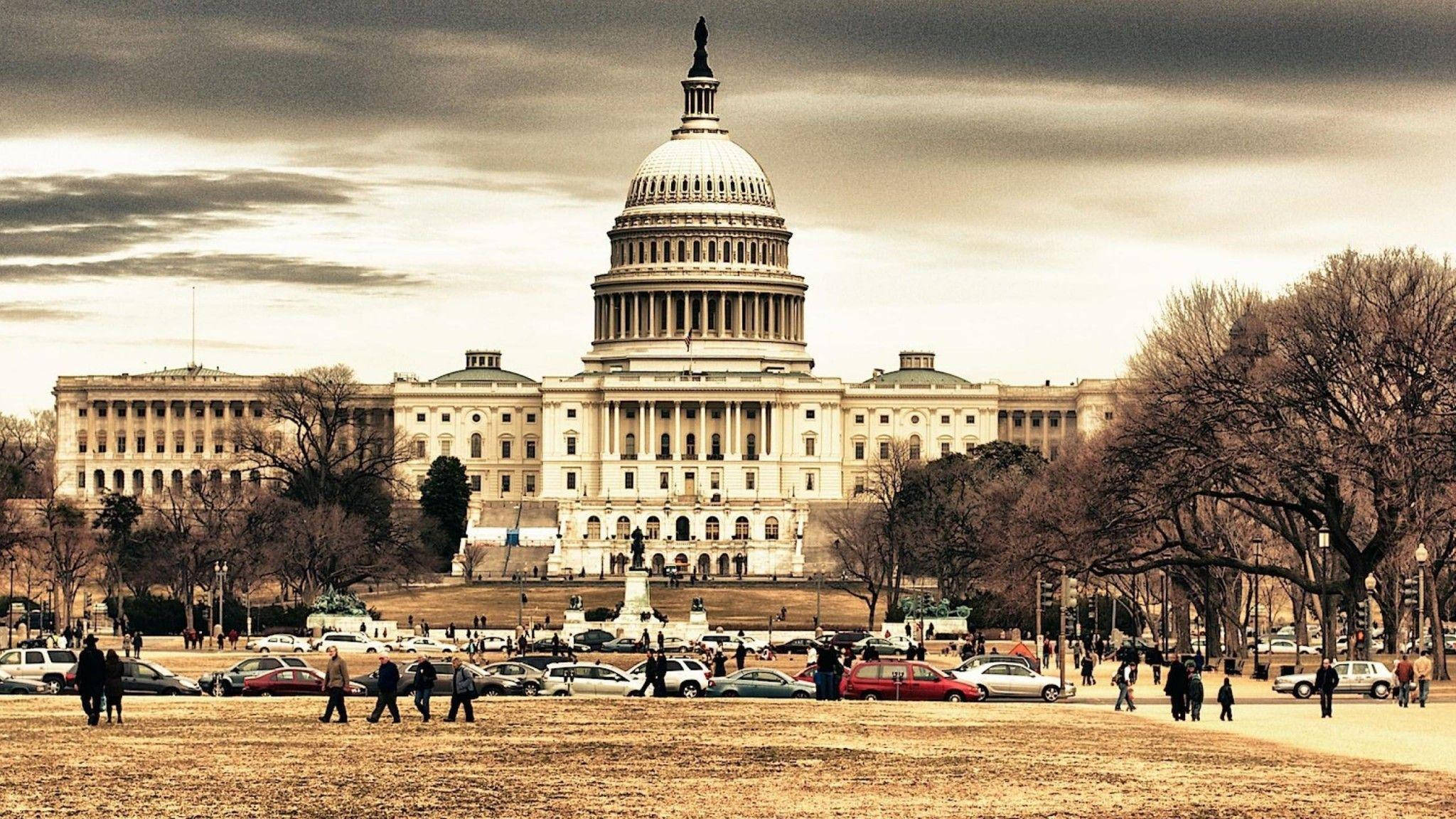 USA 桌布 壁紙 — 美國國會大廈在戲劇性的多雲天空下巍然聳立，周圍有光禿禿的樹木和人們走過草坪。