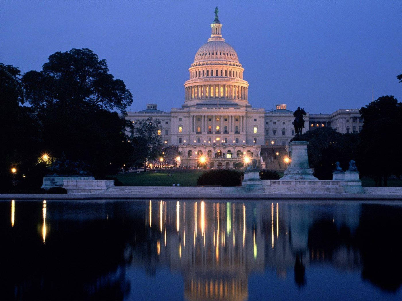Washington 桌布 壁紙 — 美國國會大廈在黃昏時溫暖地發光，其圓頂和窗戶倒映在一個由雕像和樹木環繞的寧靜水池中。
