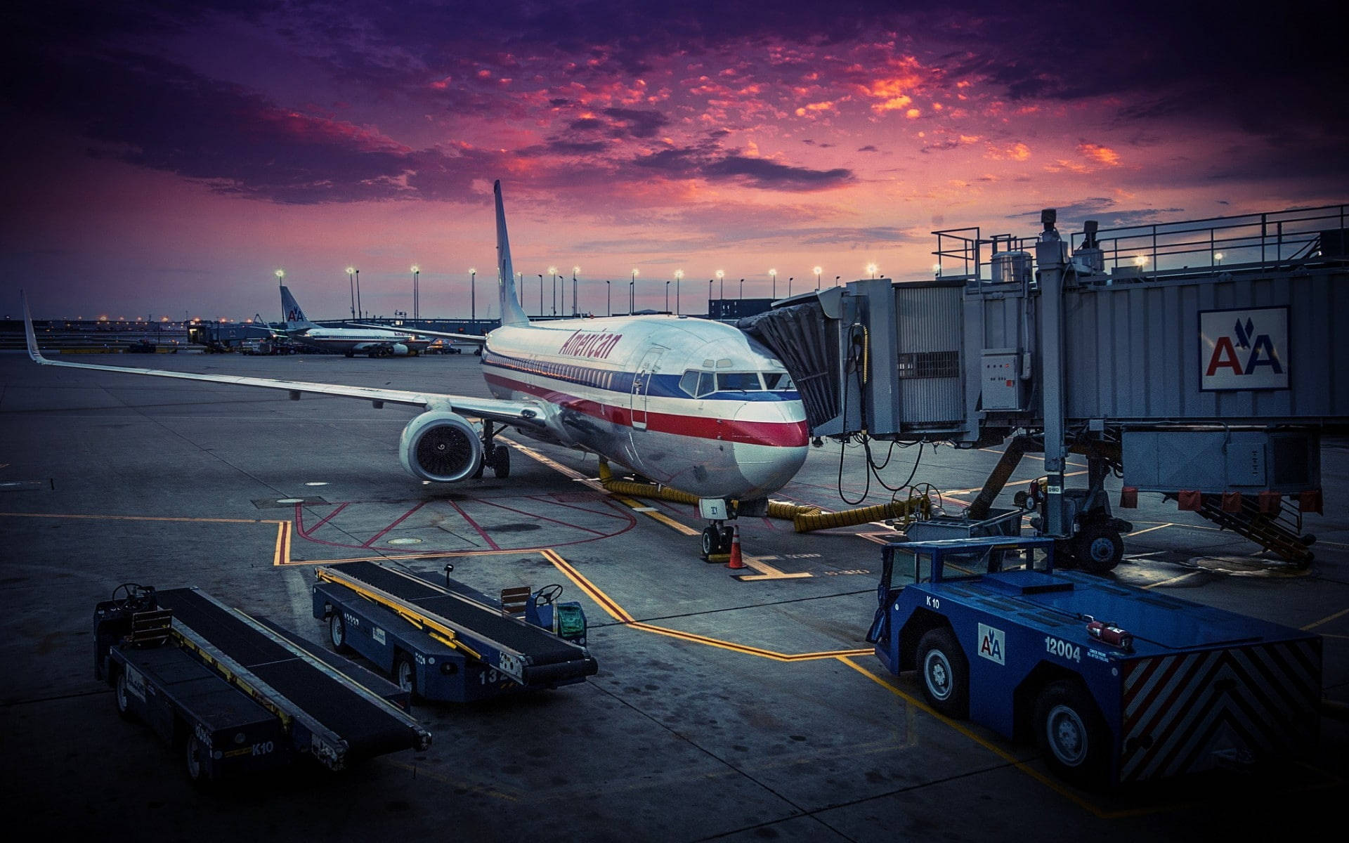 HD Plane 桌布 壁紙 — 美國航空噴射機停在登機門，背景是戲劇性的紫橙色暮色天空，附近有機場地面設備。
