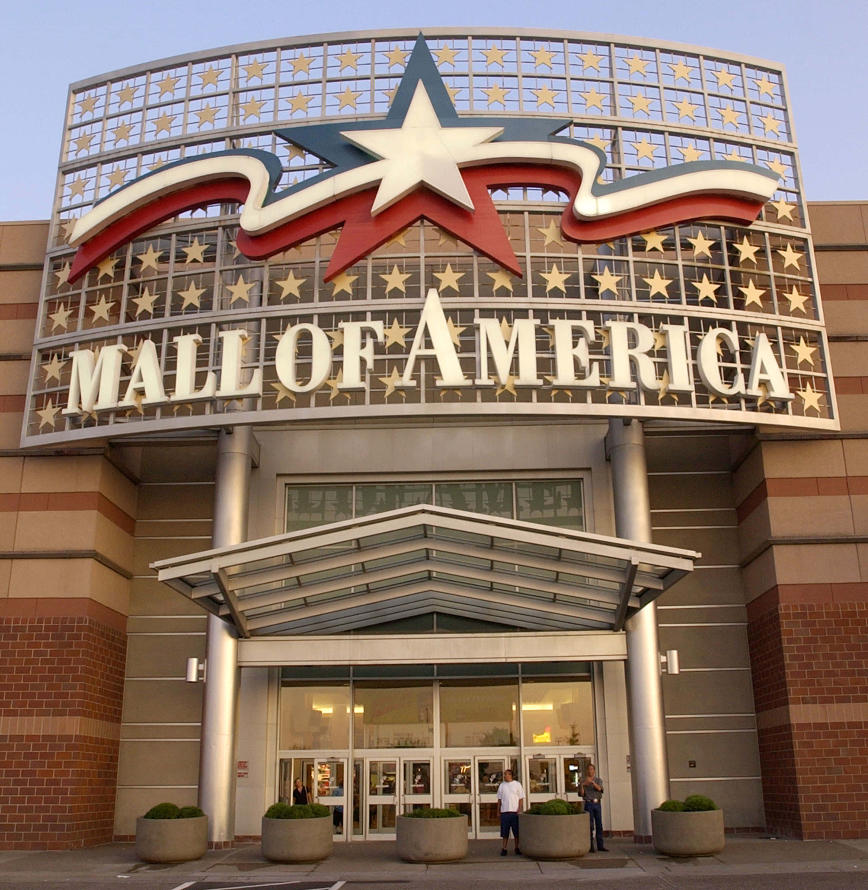 Mall Of America 桌布 壁紙 — 美國購物中心宏偉的入口處，在晴朗的天空下有一個巨大的愛國星形和絲帶標誌，人們站在玻璃門附近。