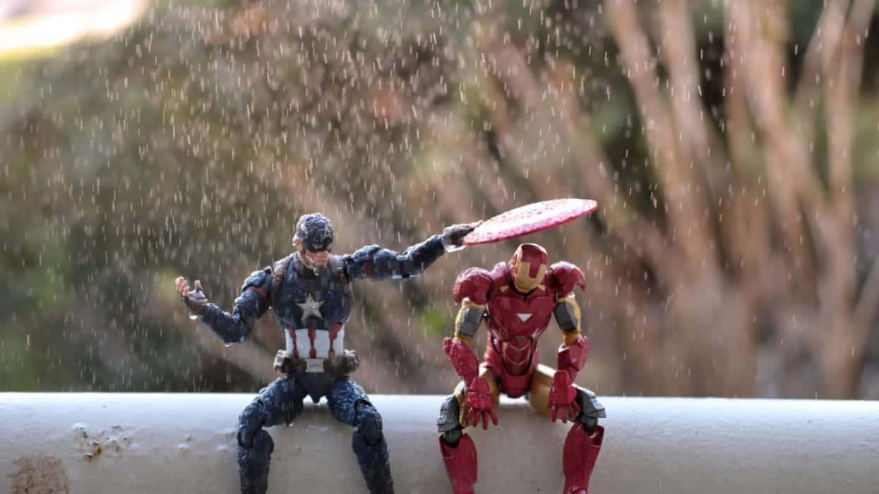 Iron Man Action Figures 桌布 壁紙 — 美國隊長和鋼鐵人的動作人偶並排坐在壁架上，雨滴在模糊的自然背景中落在它們周圍。