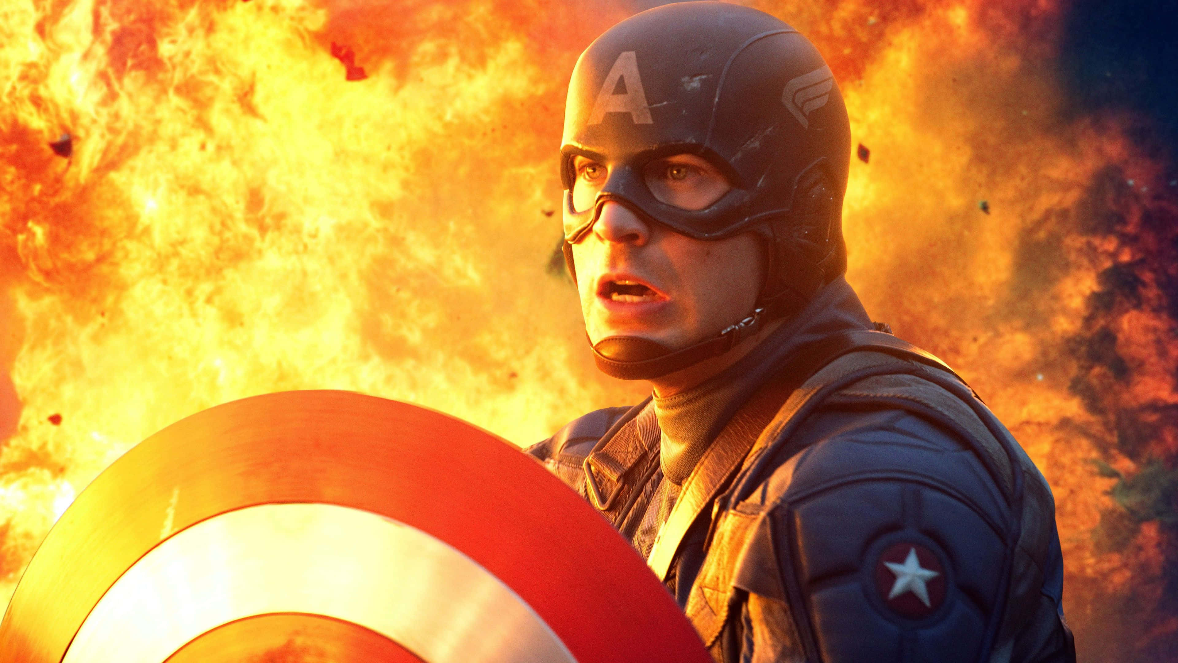 Captain America Desktop 桌布 壁紙 — 美國隊長在洶湧的橙色火焰和煙霧背景下堅定地舉著盾牌。