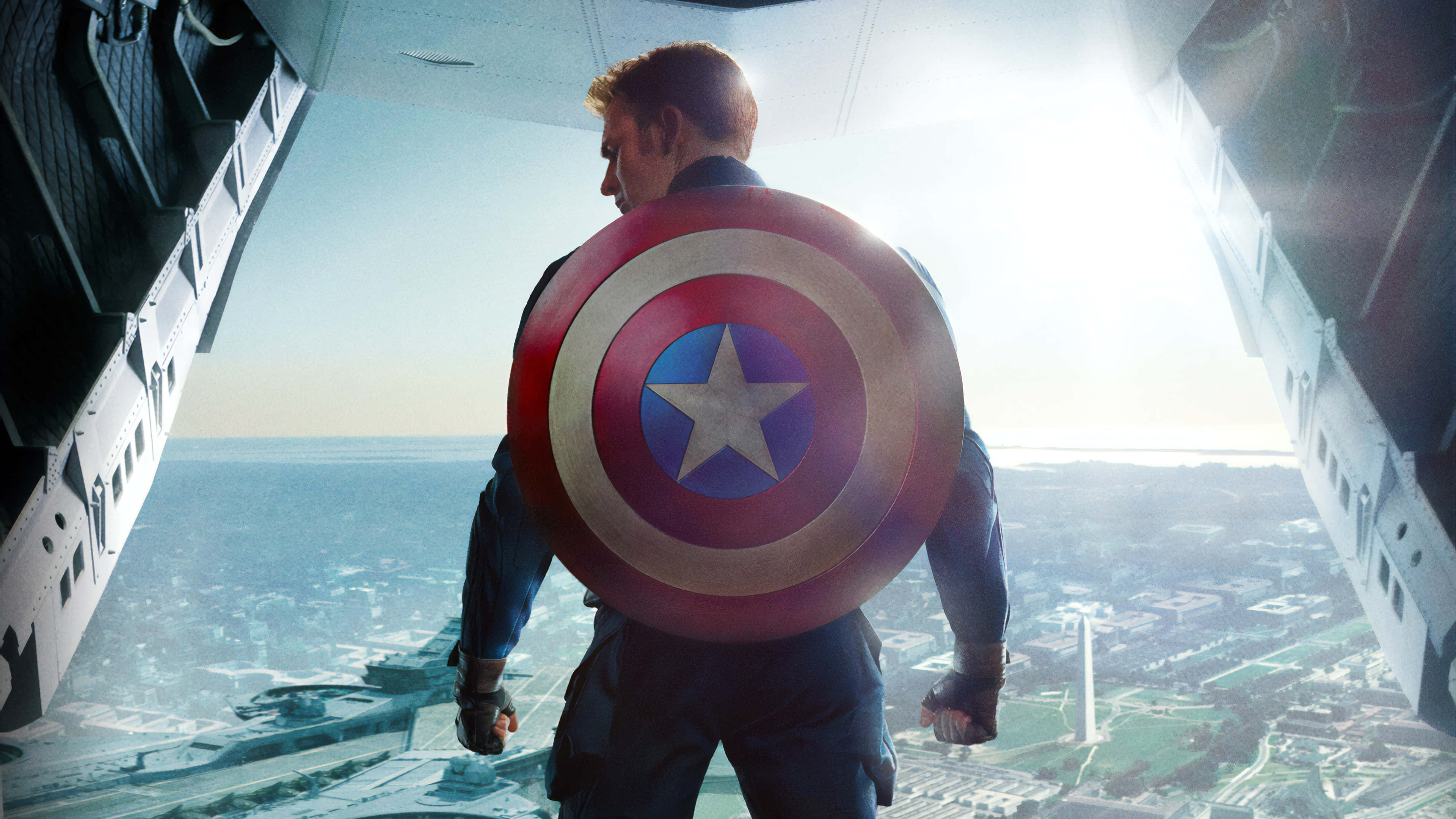 Captain America Desktop 桌布 壁紙 — 美國隊長站在飛機的敞開門口，手持盾牌，俯瞰著沐浴在明亮日光下的廣闊城市景觀。