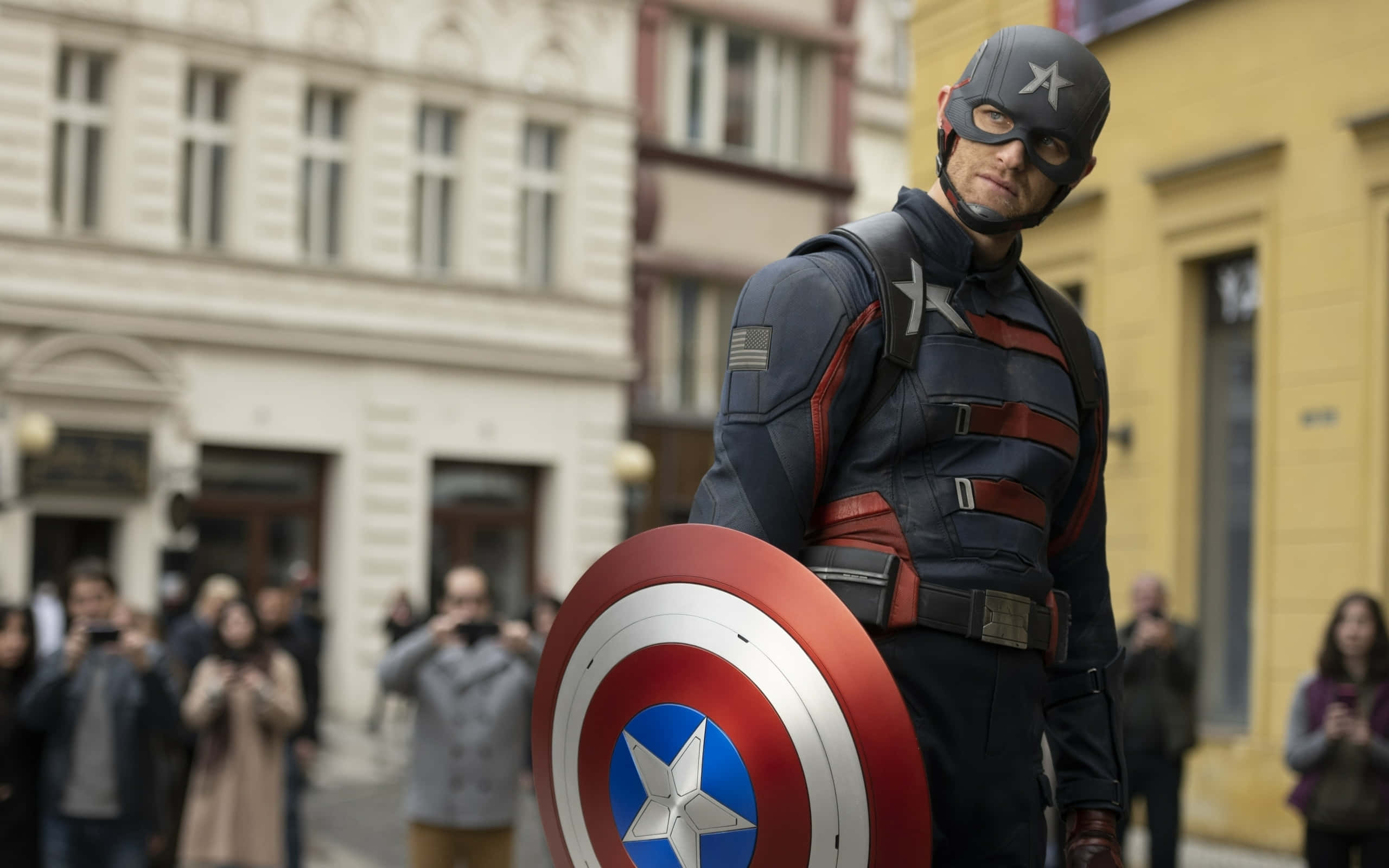 Captain America Movie 桌布 壁紙 — 美國隊長自信地站在歐洲城市街道上，手持他標誌性的紅白藍盾牌，旁觀者正在拍攝他。