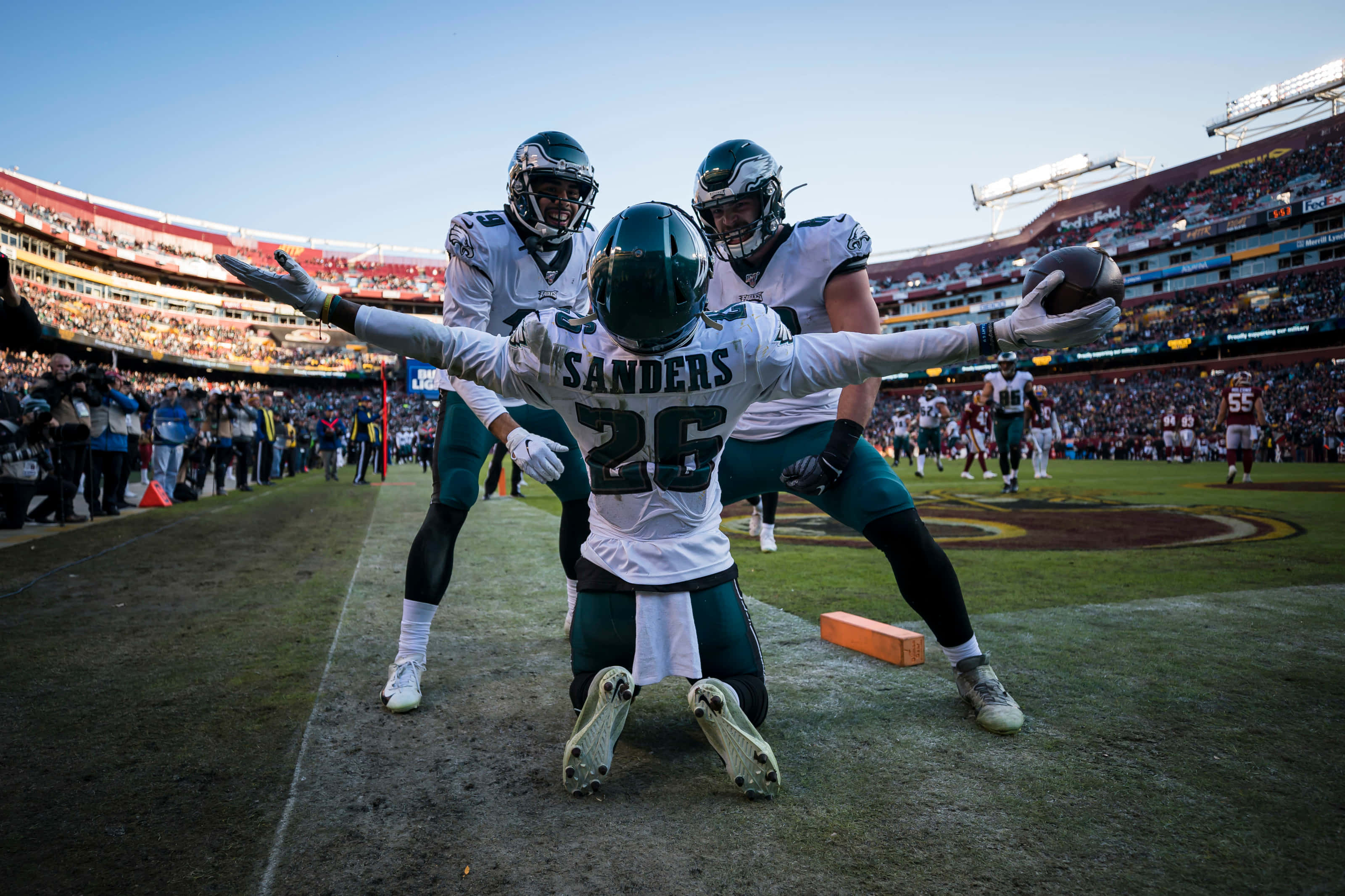 Football Players 桌布 壁紙 — 美式足球運動員桑德斯26號在球場上跪地慶祝，手持球，隊友在他身後於體育場燈光下慶祝。