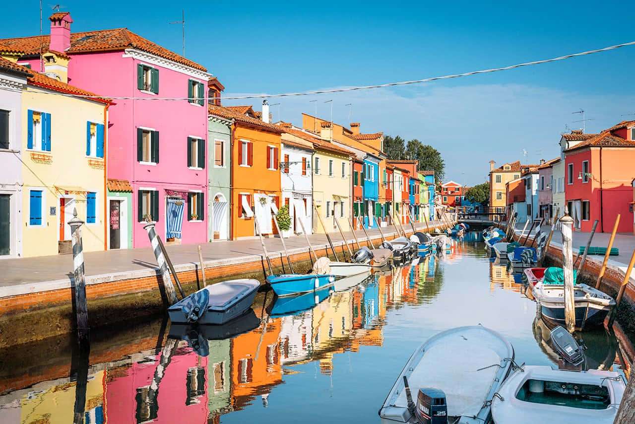 Italian Island 桌布 壁紙 — 義大利布拉諾島寧靜的運河兩旁排列著色彩繽紛的粉彩房屋，船隻停泊，清澈的藍天倒映在水中。