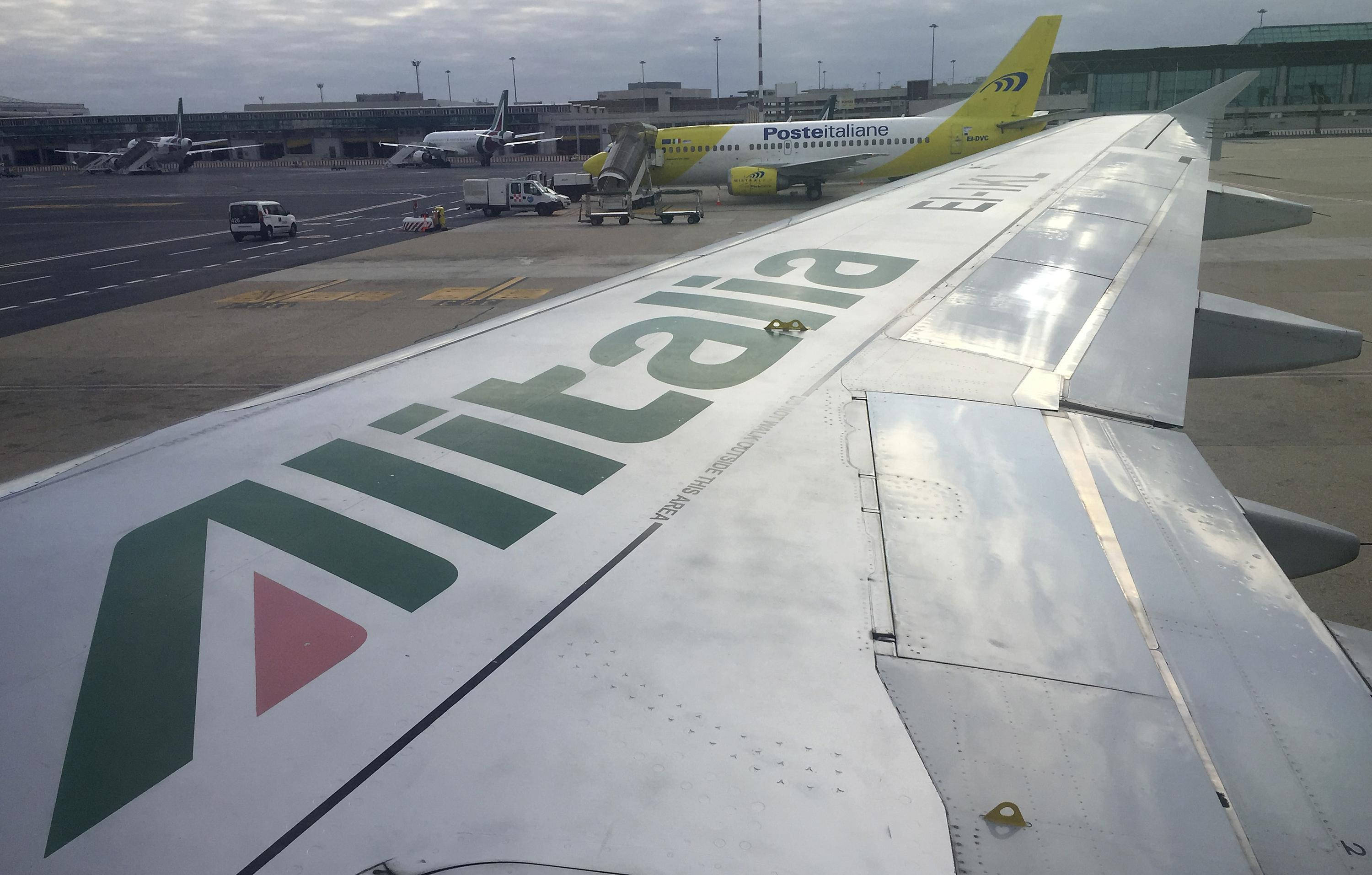 桌布 壁紙 — 義大利航空飛機機翼的特寫視圖，帶有航空公司標誌，俯瞰繁忙的機場停機坪，其他飛機和地面車輛在陰天天空下。