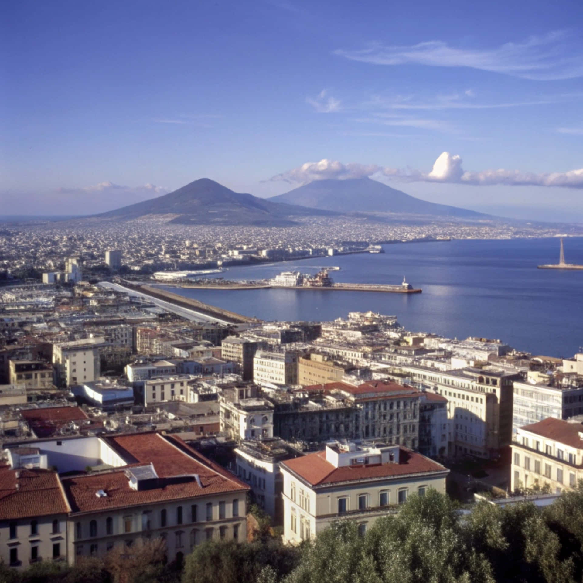 Naples skyline 壁紙 — 義大利那不勒斯的鳥瞰圖，紅瓦屋頂、港口和遠處的維蘇威火山在清澈的藍天下。