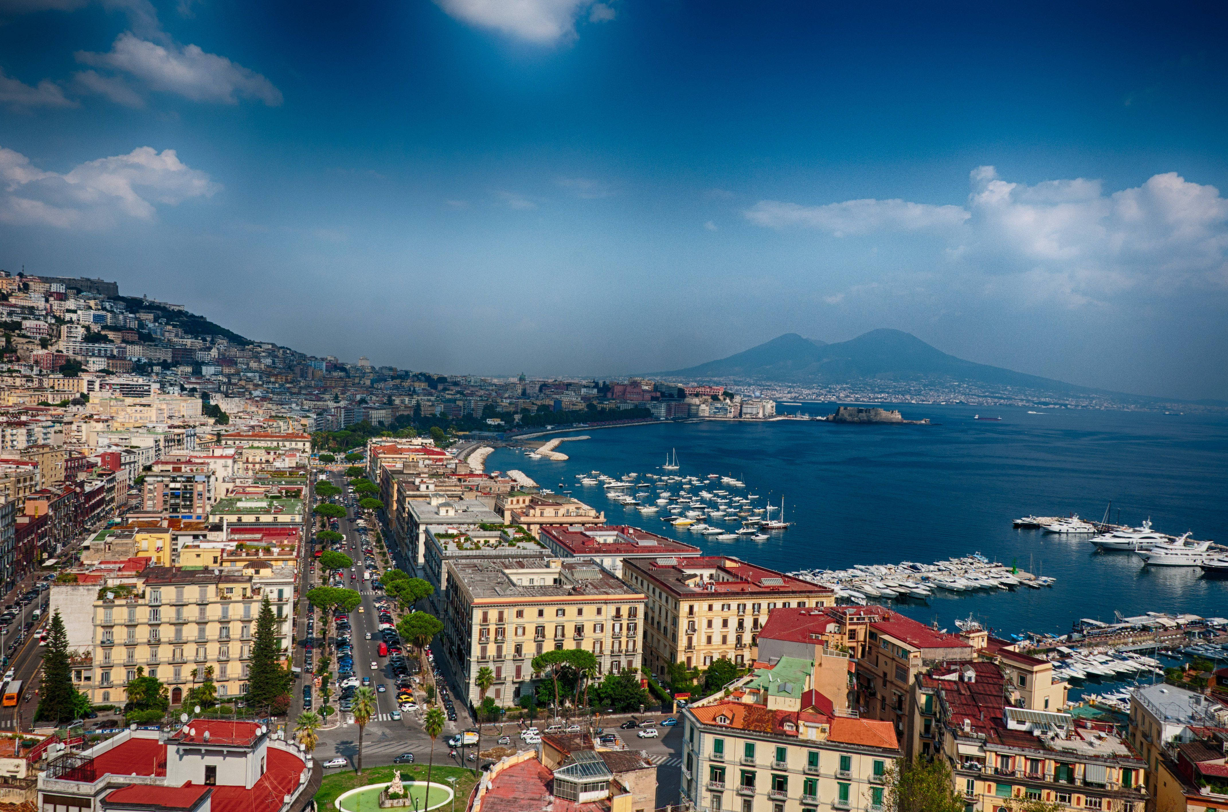 Naples 桌布 壁紙 — 義大利那不勒斯的鳥瞰圖，色彩繽紛的建築物緊貼海岸線，遠處的維蘇威火山在明亮的藍天下若隱若現。