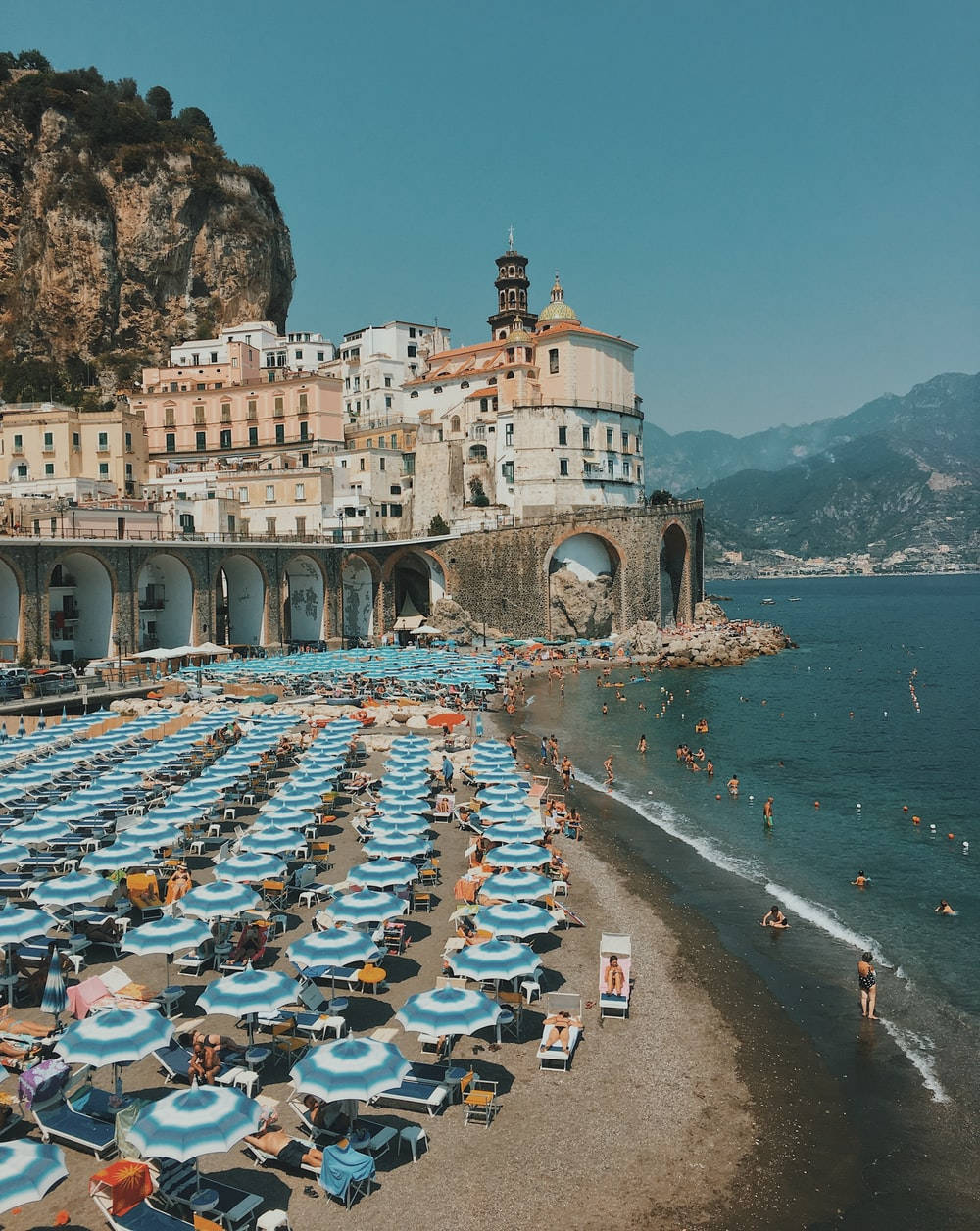 Capri 桌布 壁紙 — 義大利阿馬爾菲海岸陽光普照的海灘，排列著藍色遮陽傘，懸崖邊的歷史建築俯瞰著碧綠的海水。