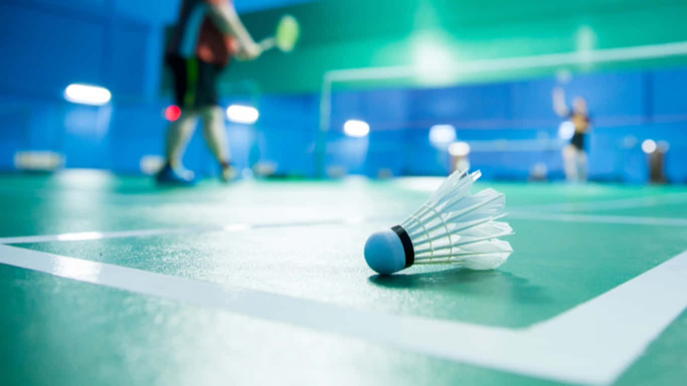 1366x768 Badminton 桌布 壁紙 — 羽毛球在綠色羽毛球場上的特寫，背景有模糊的運動員在移動。