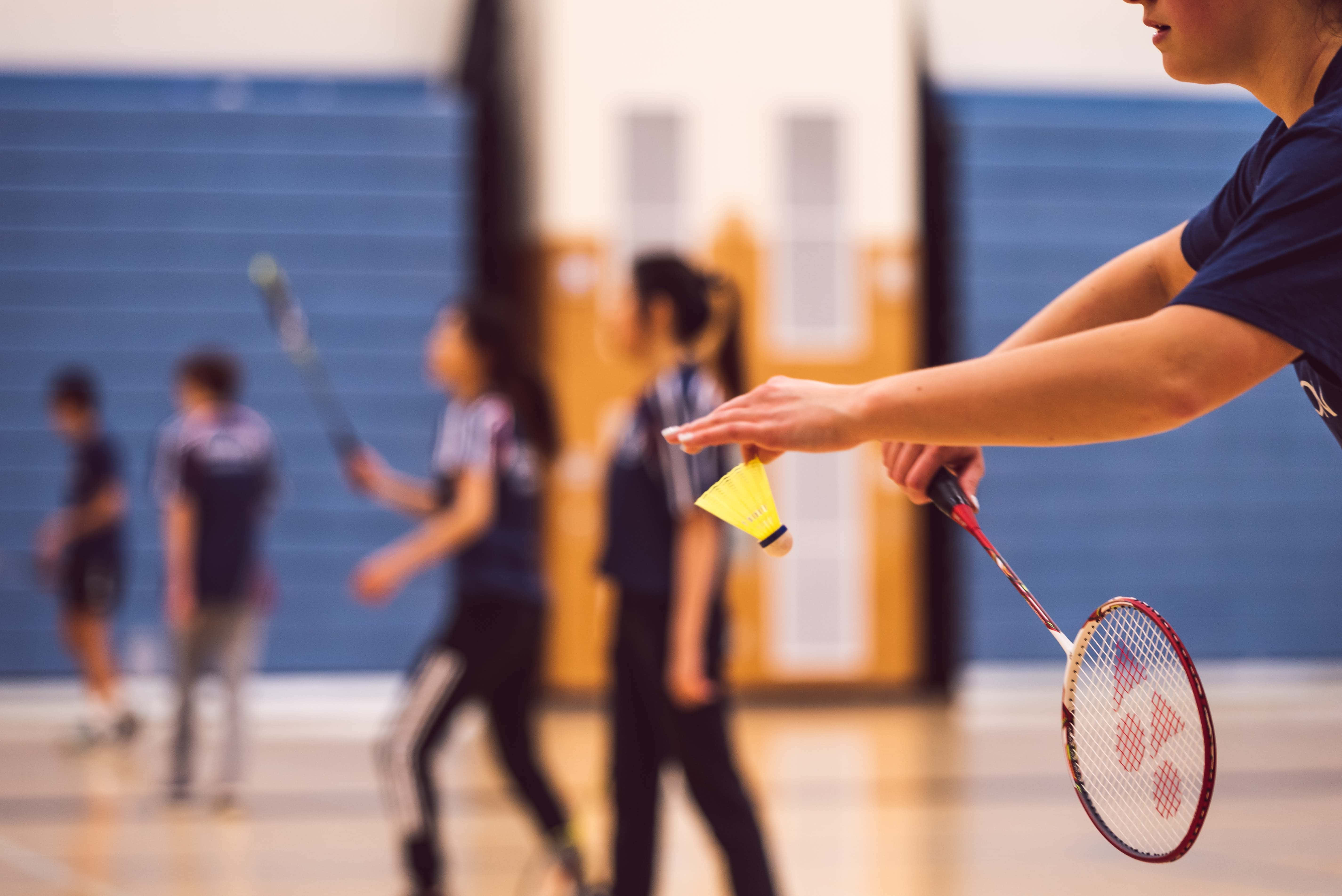 Badminton 桌布 壁紙 — 羽毛球運動員手持球拍和羽毛球的特寫，背景中隊友模糊，在室內場地上。
