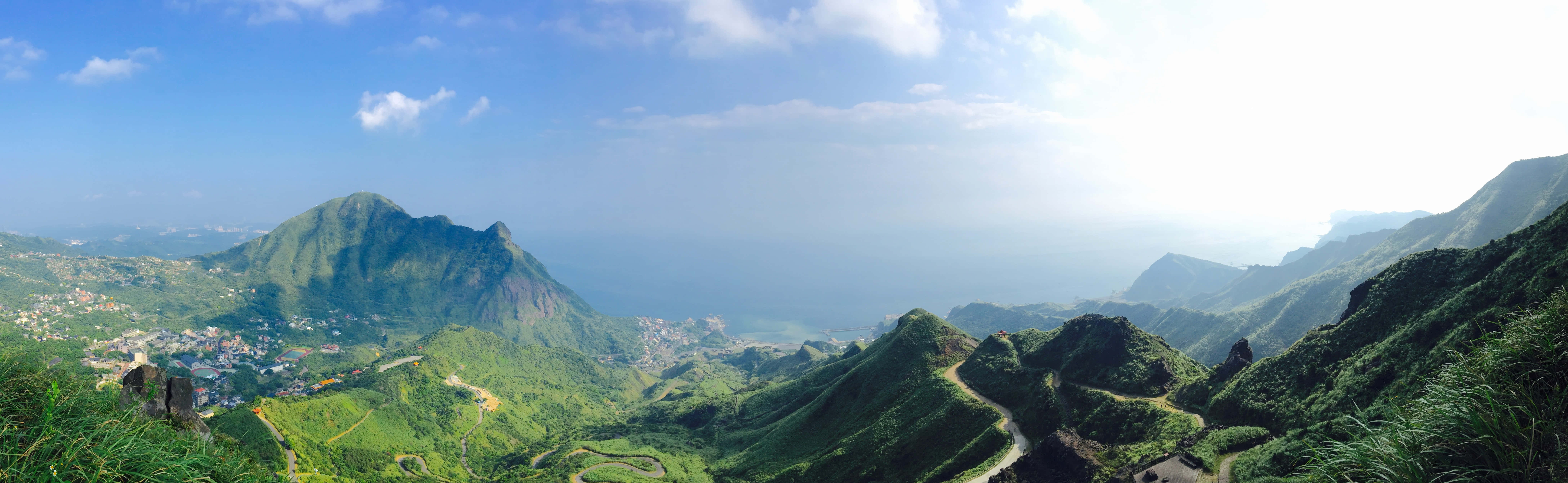 Minimalist Dual Monitor 桌布 壁紙 — 翠綠山巒的廣闊全景，蜿蜒的道路和沿海小鎮，在明亮的藍天下點綴著幾朵白雲。