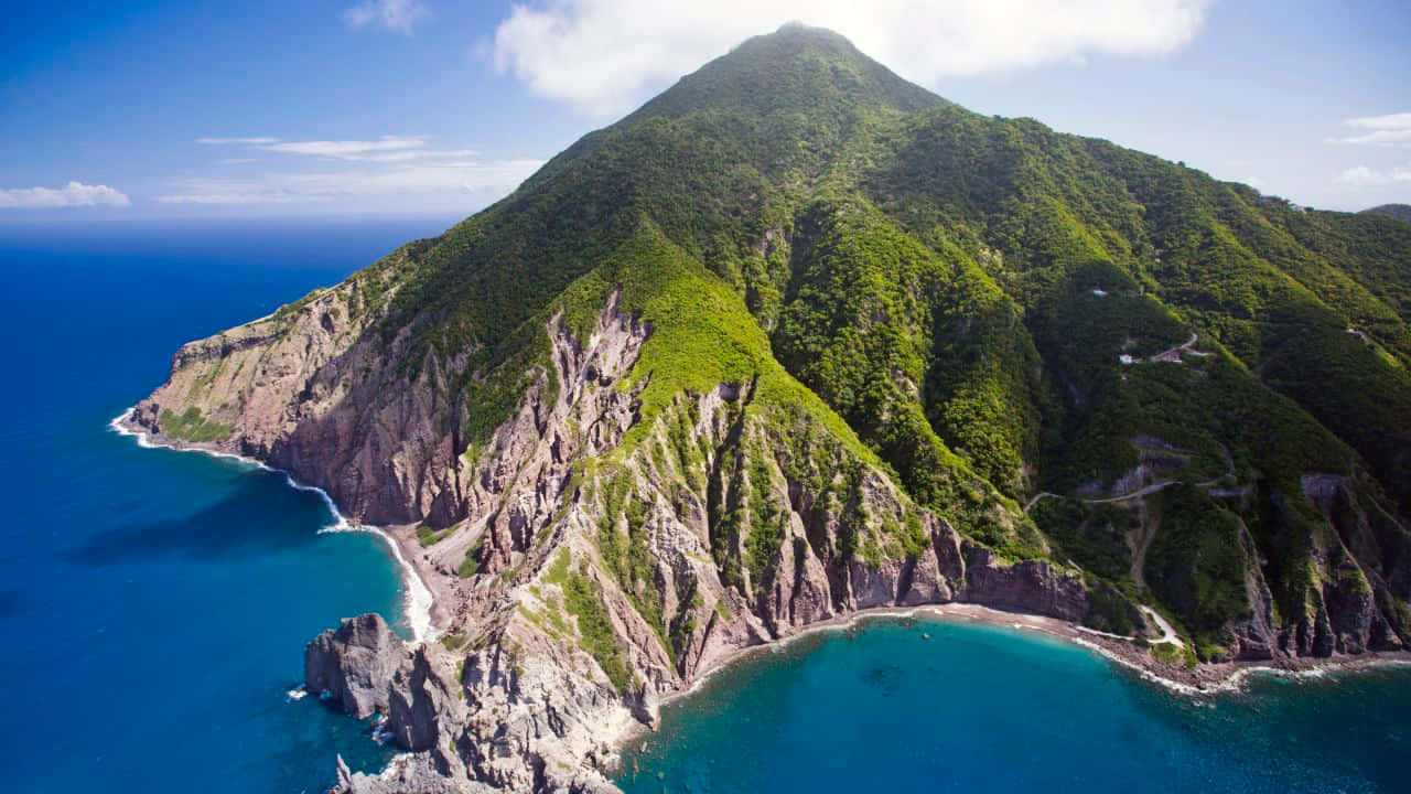 Caribbean Island 桌布 壁紙 — 翠綠山巒突出於充滿活力的藍綠色海洋，蜿蜒的海岸道路的鳥瞰圖。