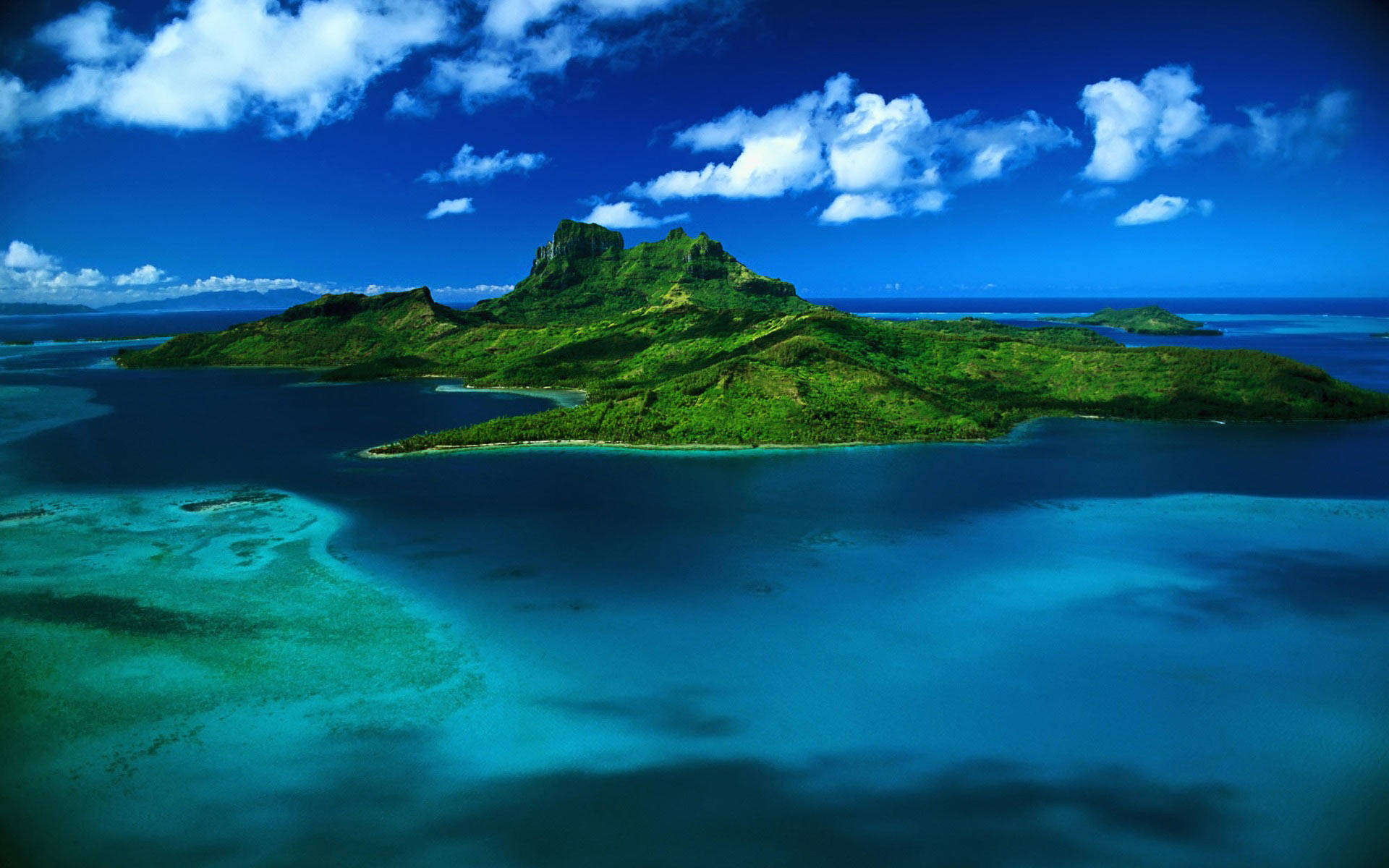 Caribbean 桌布 壁紙 — 翠綠島嶼的鳥瞰圖，島上有火山峰，周圍環繞著充滿活力的綠松石色潟湖和深藍色海洋，天空明亮，有蓬鬆的白雲。