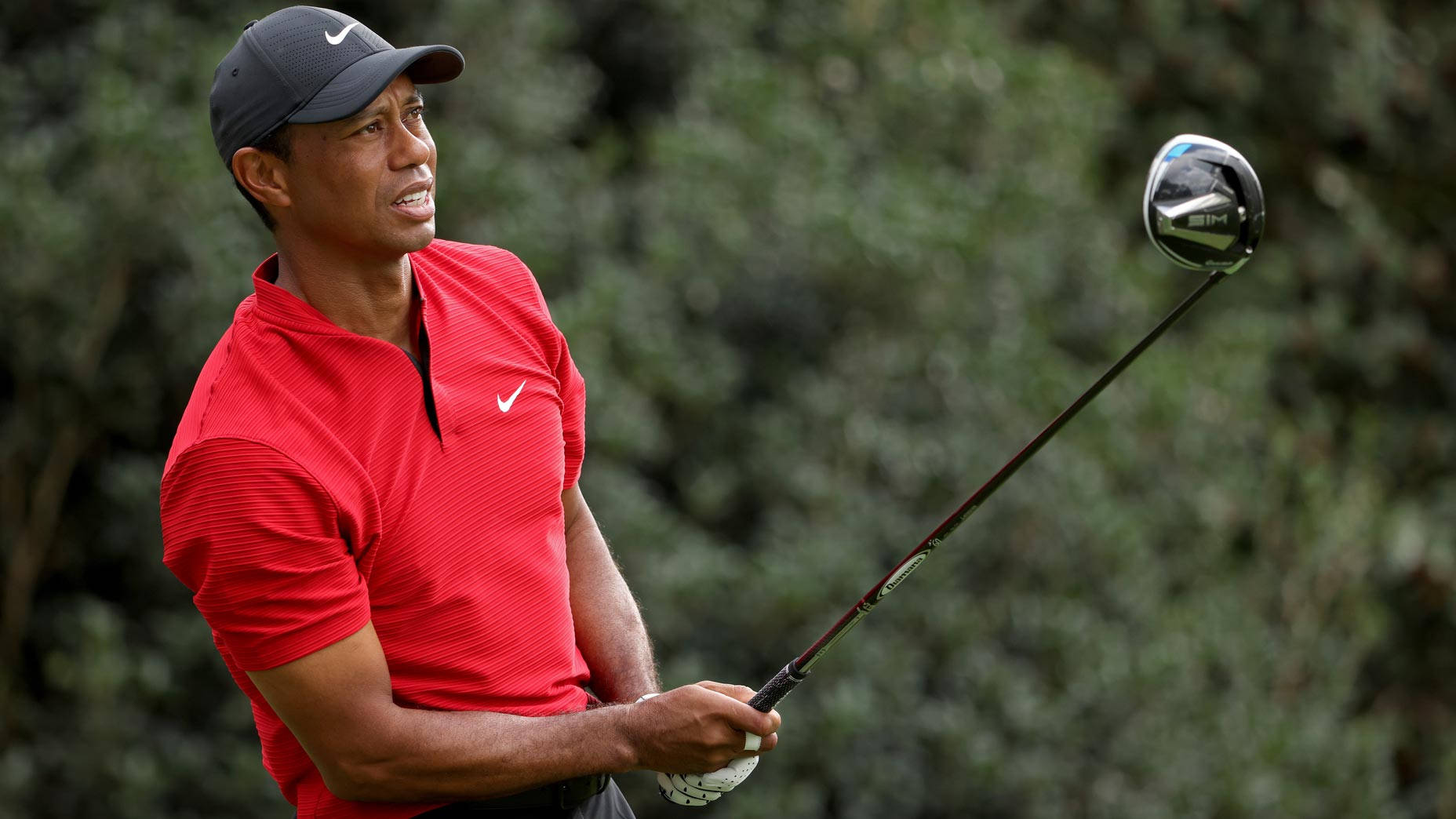 woods 桌布 壁紙 — 老虎伍茲身穿紅色Nike Polo衫和帽子，在模糊的綠色樹葉背景下揮桿。