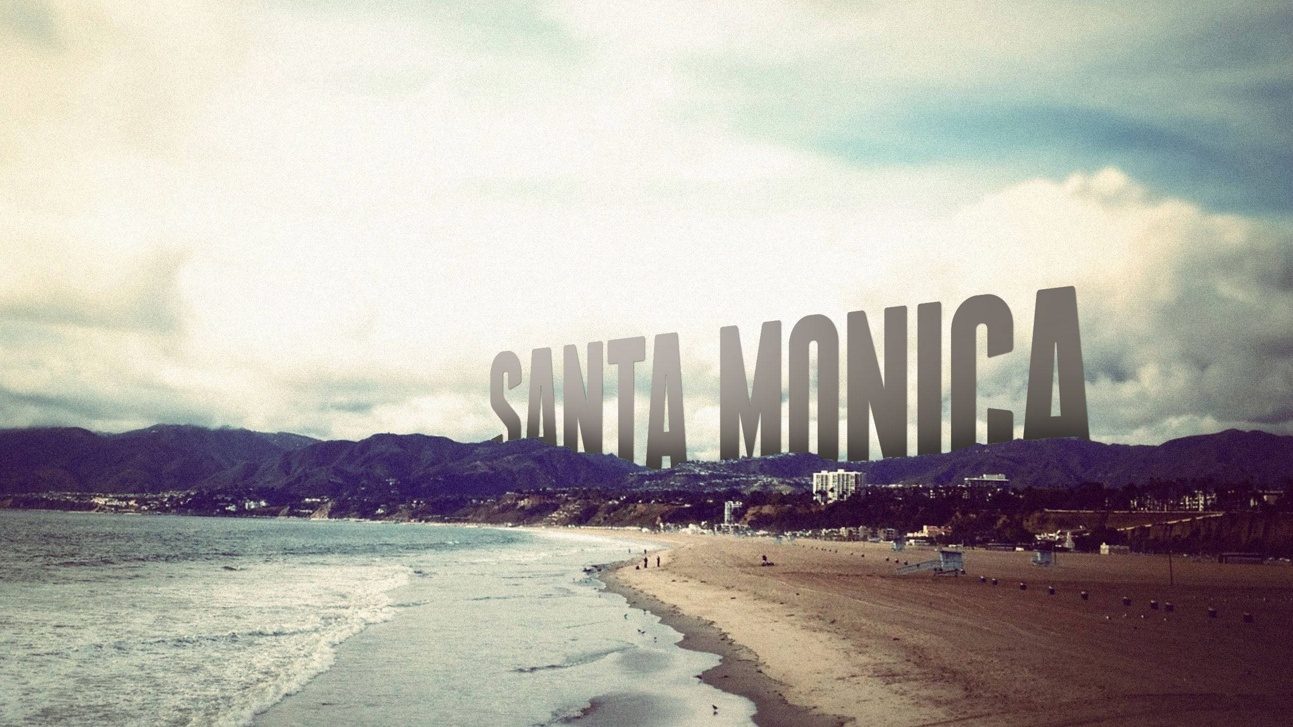 Monica 桌布 壁紙 — 聖塔莫尼卡標誌聳立在多雲的海灘上，海浪拍打，遠山在暗淡的天空下若隱若現。