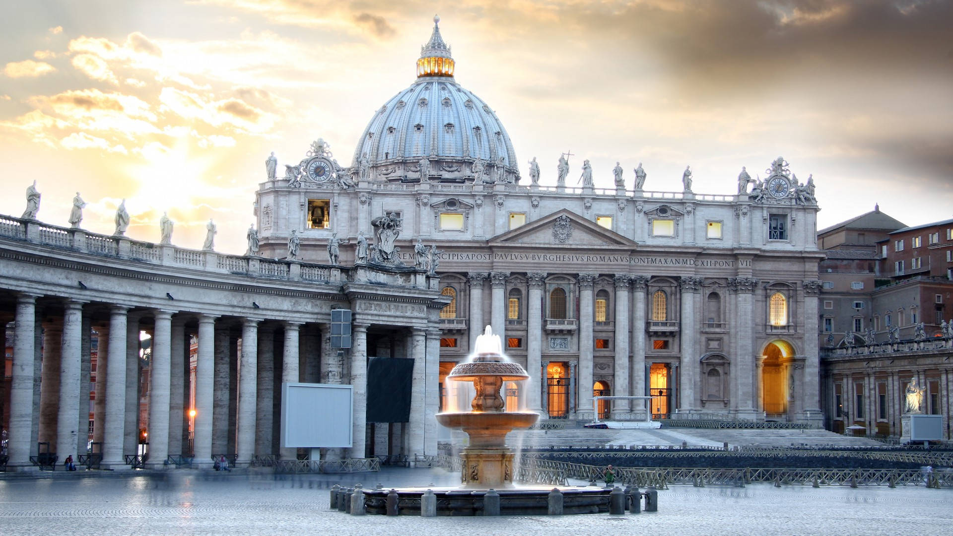 Vatican 桌布 壁紙 — 聖彼得大教堂沐浴在金色日落光芒中，其標誌性圓頂和噴泉在濕漉漉的廣場上倒映。