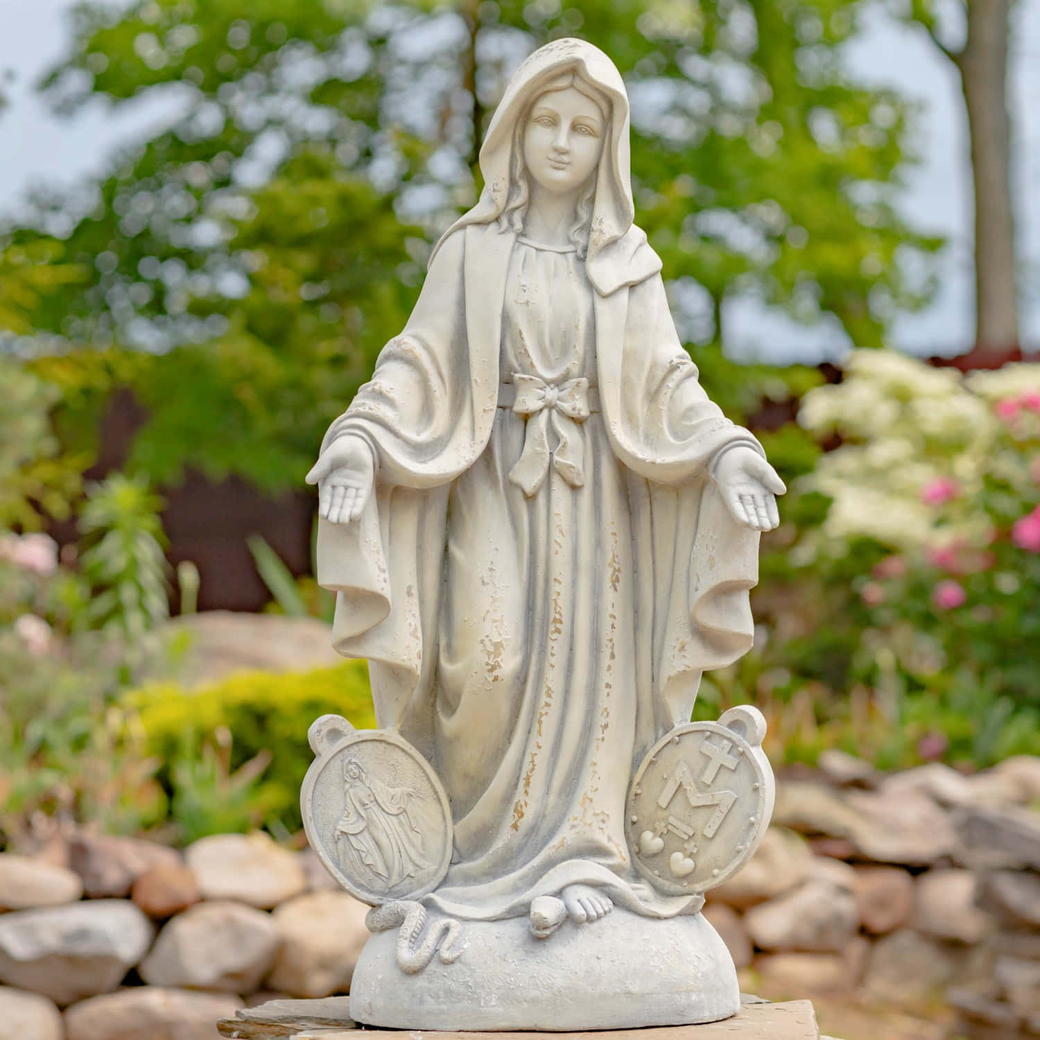 Mother Mary 桌布 壁紙 — 聖母瑪利亞的白色石像優雅地矗立在茂盛的花園中，背景是柔和的綠色樹葉和粉紅色的花朵。