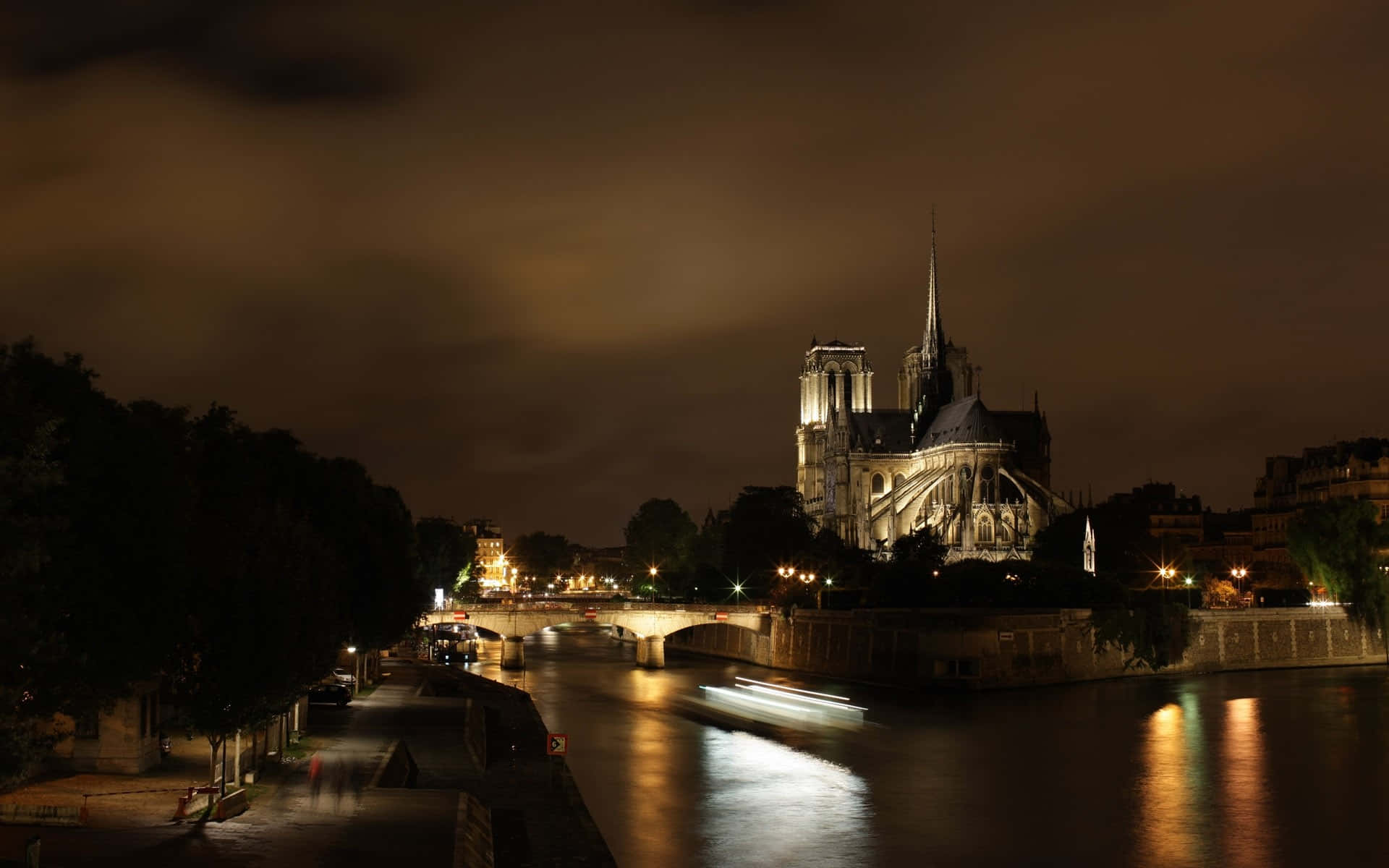Notre Dame 桌布 壁紙 — 聖母院在夜燈下溫暖地發光，倒映在塞納河上，帶有模糊的船隻軌跡。