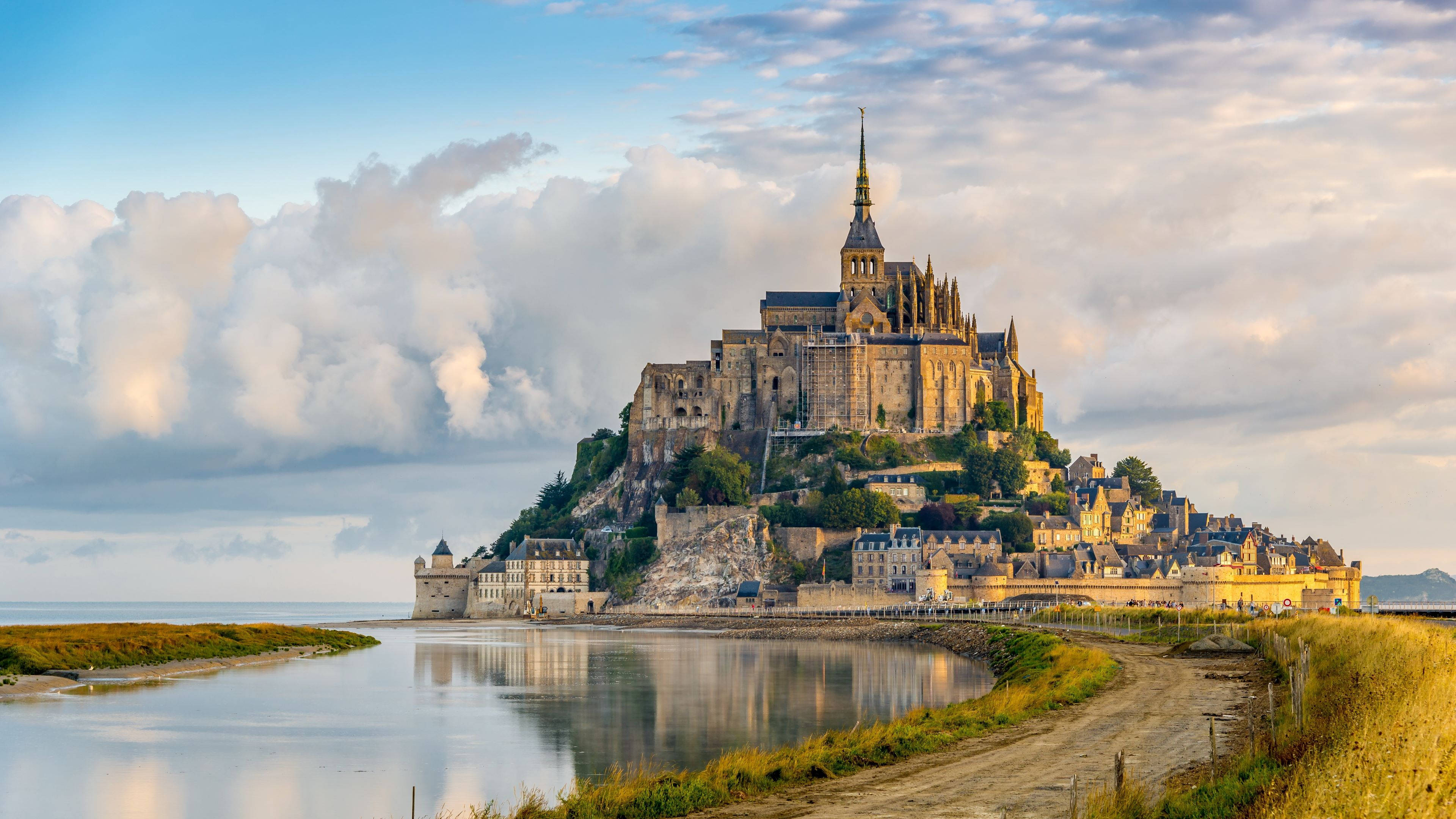 France 桌布 壁紙 — 聖米歇爾山在柔和的金色日落天空下，從潮汐水域中雄偉地升起，伴隨著蓬鬆的雲朵。