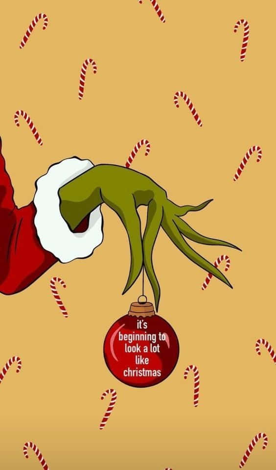桌布 壁紙 — 聖誕怪傑的綠色手在紅色袖子中拿著一個帶文字的紅色裝飾品，周圍是落在棕褐色背景上的糖果杖。
